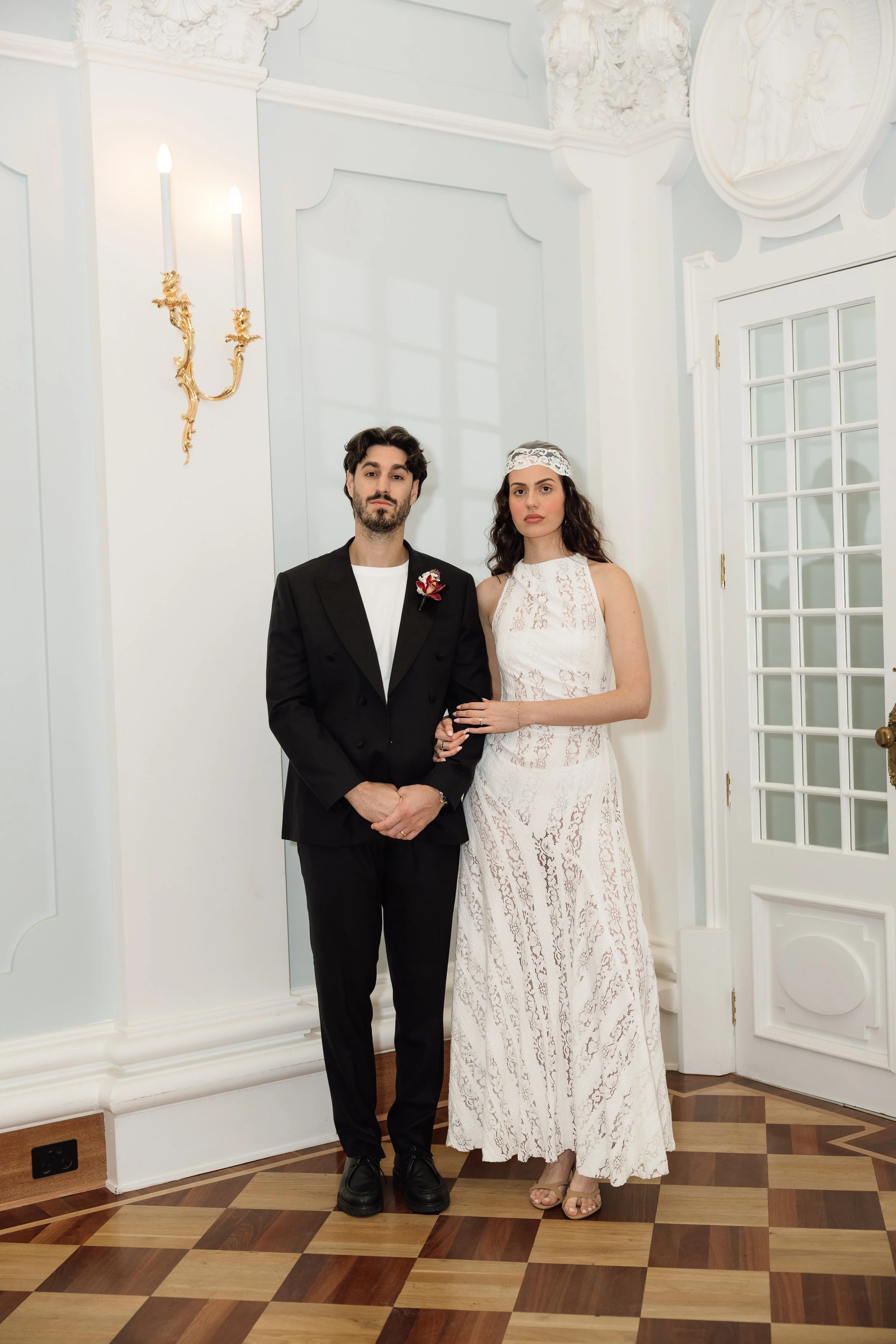 Couple posing in wedding attire inside an elegant room with ornate white molding, gold sconces, and patterned wood flooring