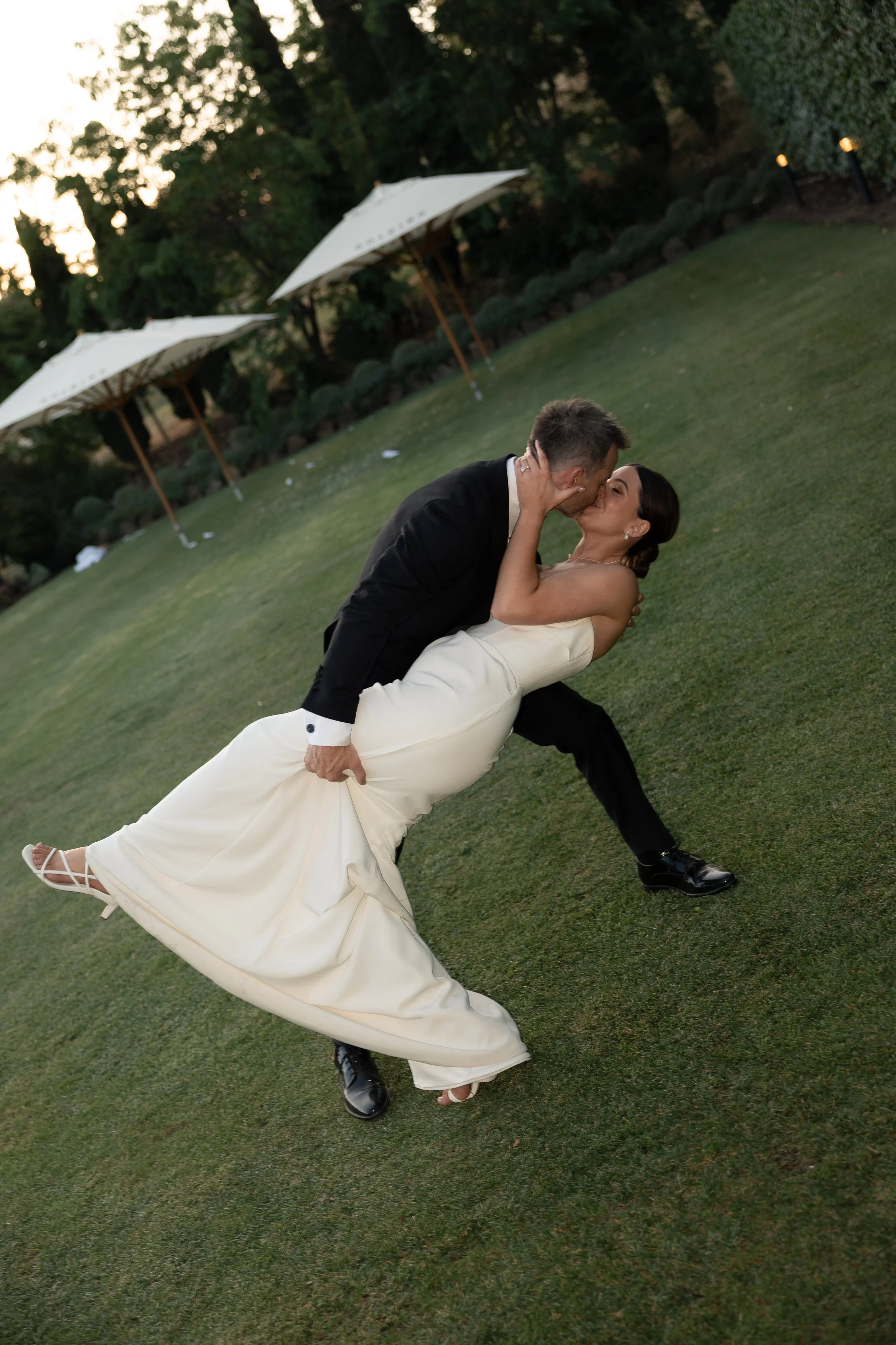 A bride and groom share a kiss on a grassy outdoor area, with umbrellas and trees in the background during sunset.