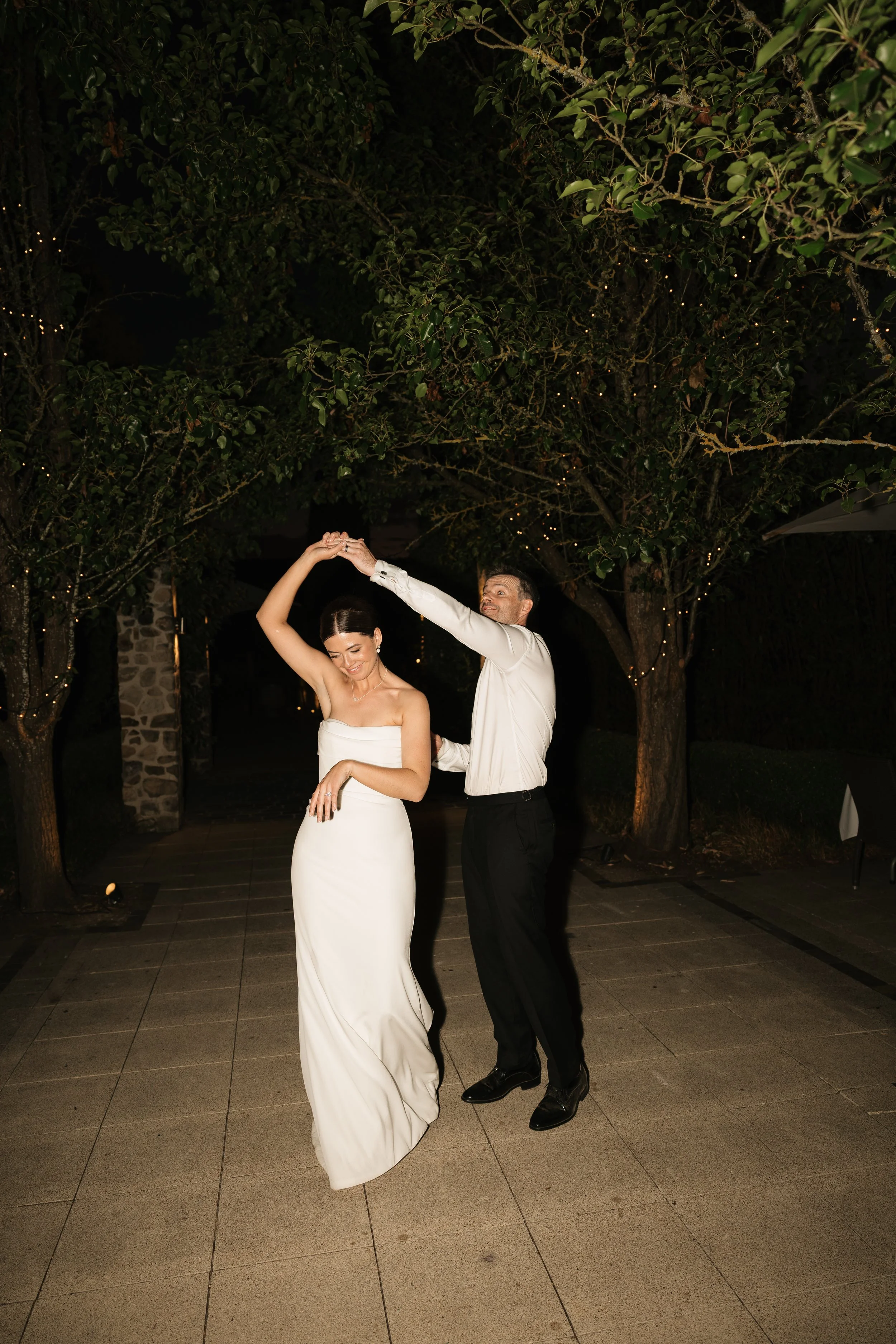 A bride and groom dancing outdoors at night, under a large tree decorated with string lights.