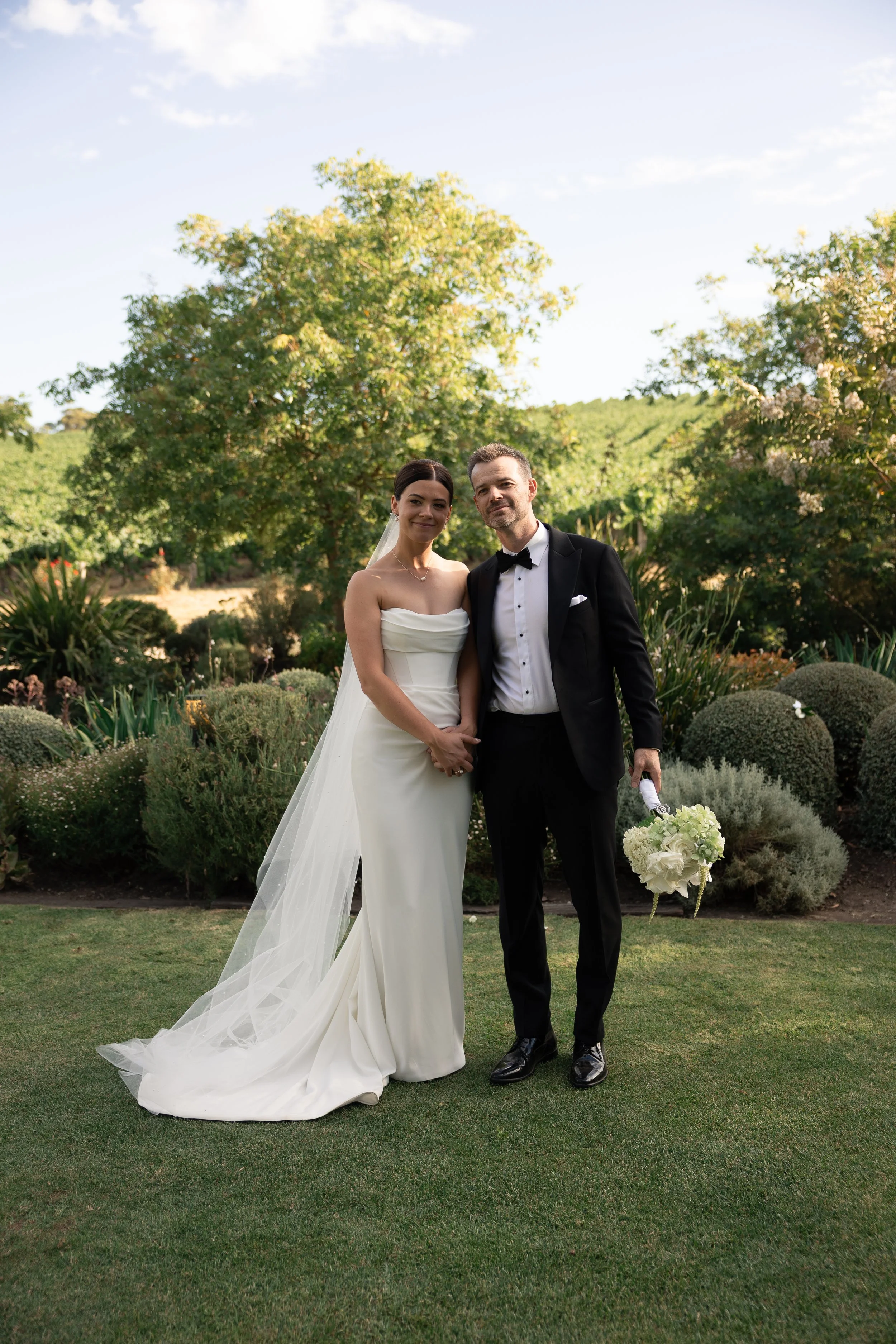 A bride and groom standing outdoors on a wedding day, holding hands, surrounded by greenery and bushes, with the bride in a white gown with a veil and the groom in a black tuxedo holding a bouquet.