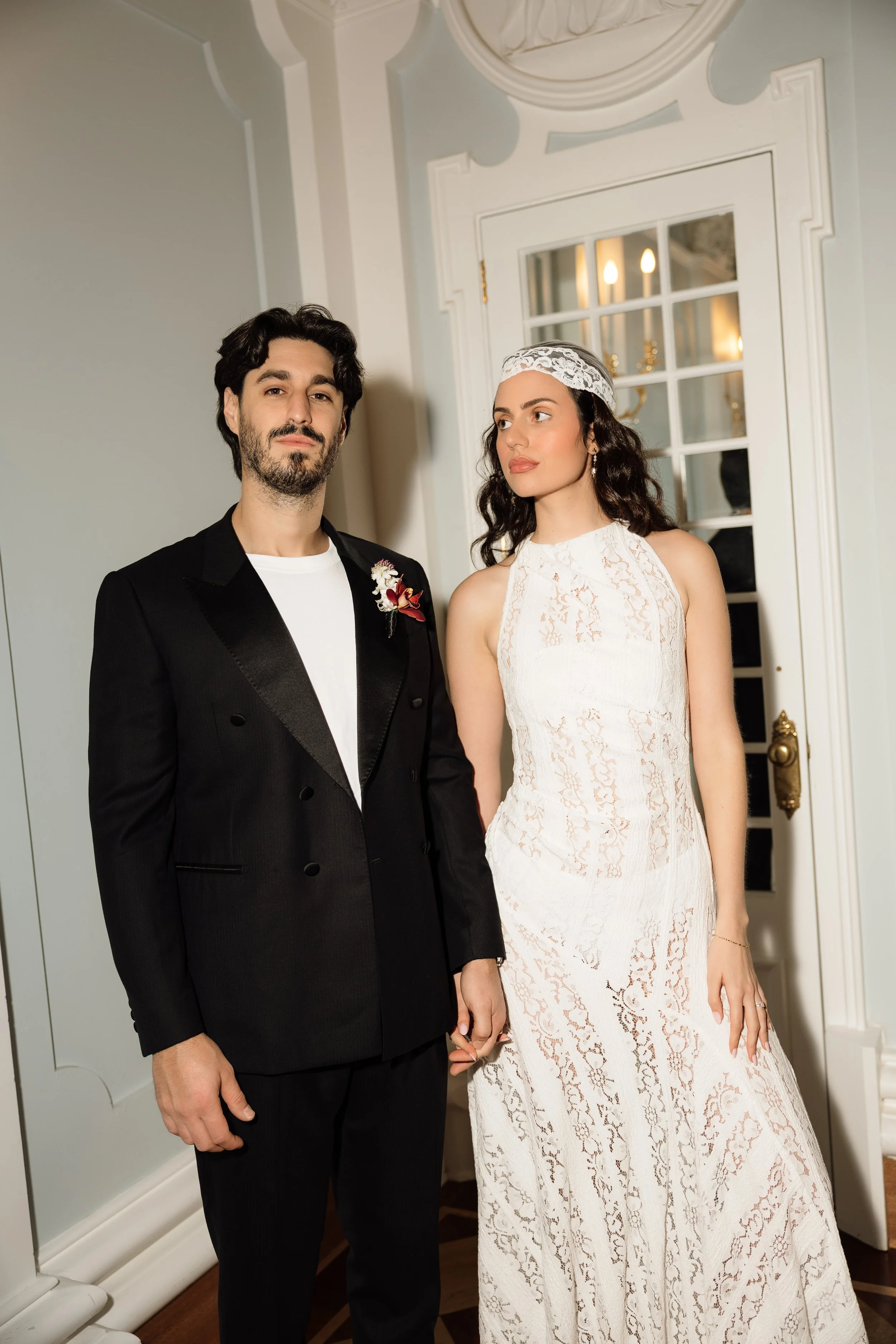 Couple posing indoors in formal attire, with the man wearing a black suit and boutonniere and the woman wearing a white lace dress and headband.