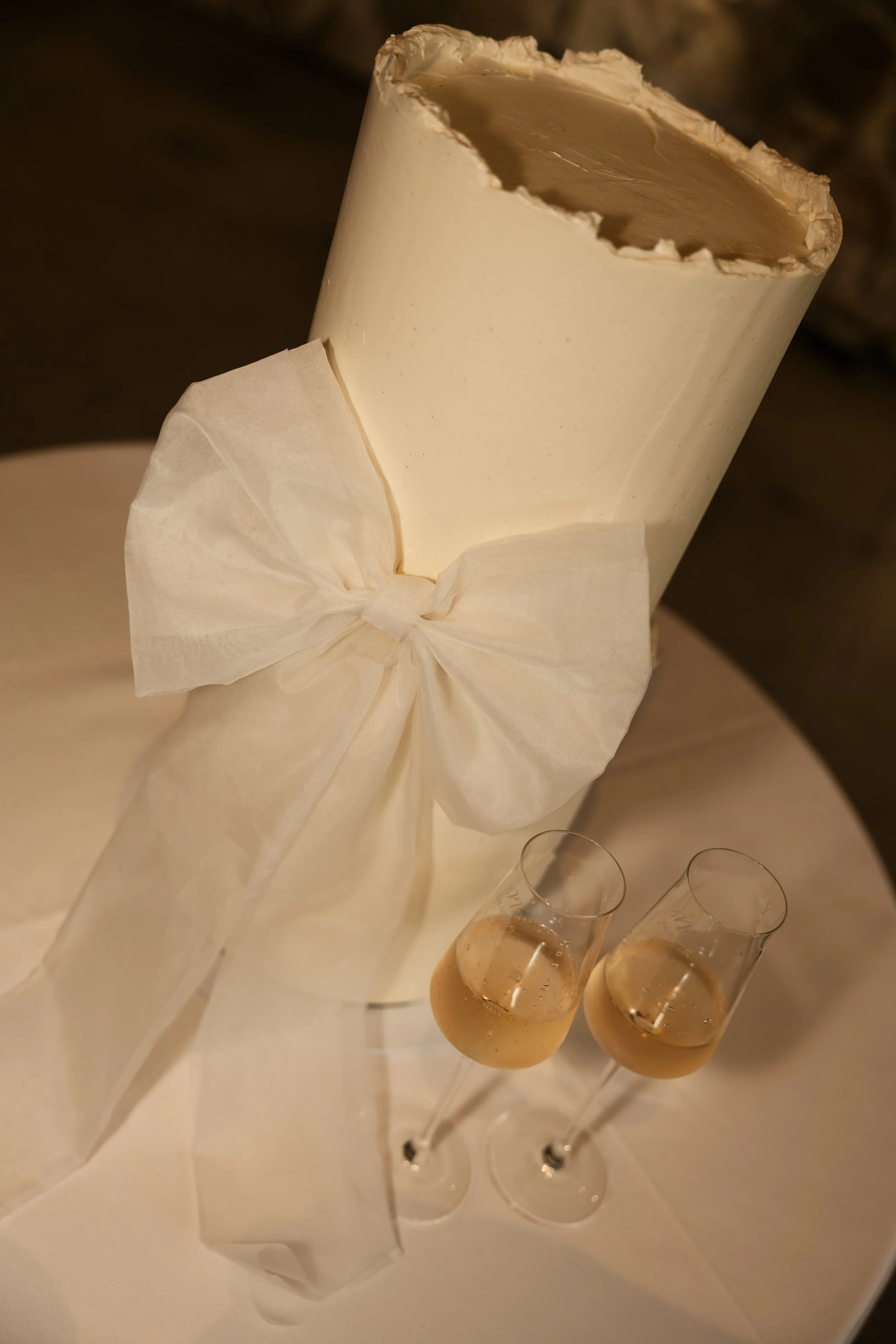 A white champagne bottle with a large bow tied around it, placed on a white surface. Two filled champagne flutes are positioned in front of the bottle.