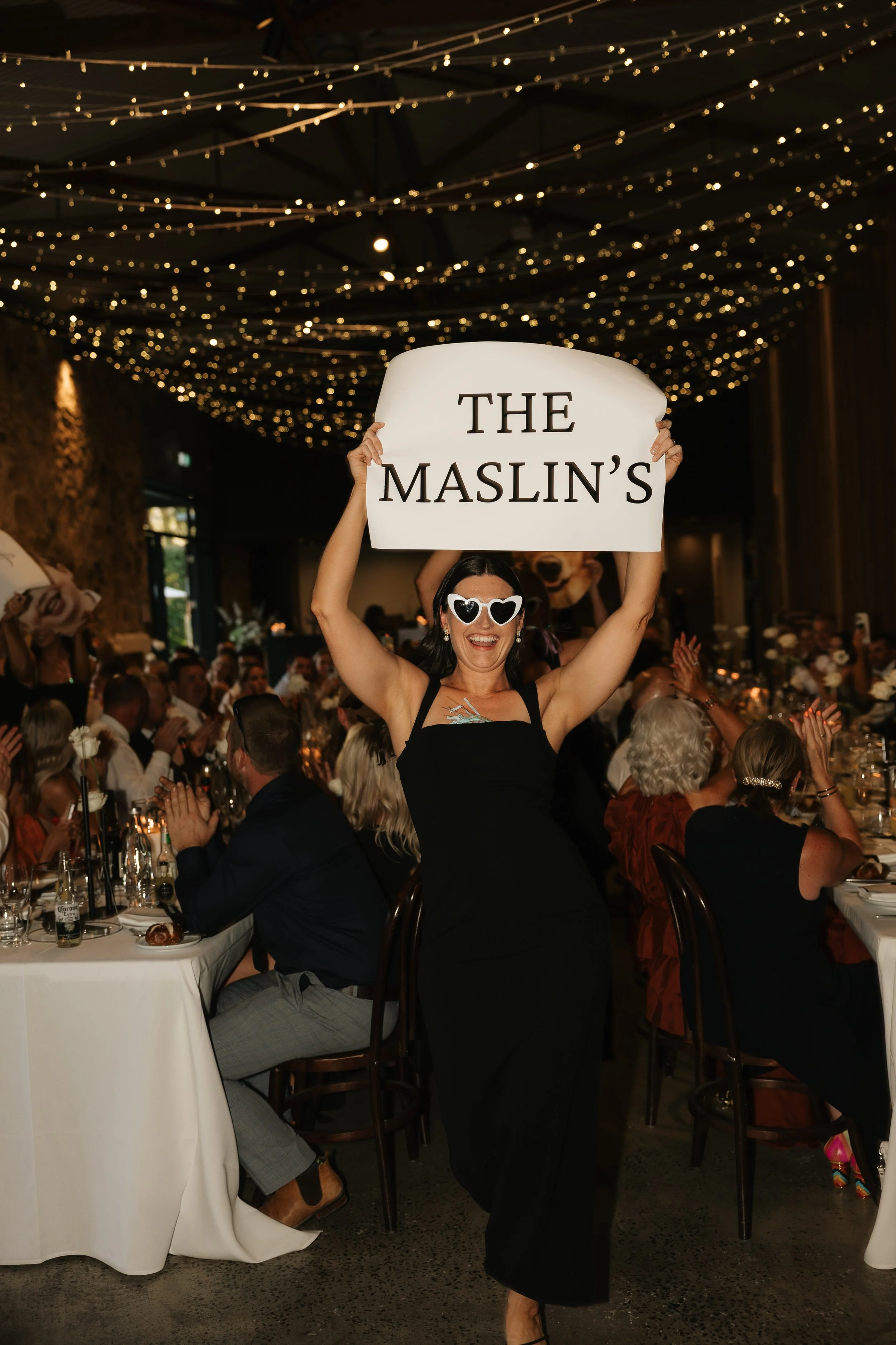 A woman wearing heart-shaped sunglasses and a black dress holding a sign that reads "THE MASLIN'S" at a formal event with many guests seated at banquet tables, string lights hanging from the ceiling, and a large dog head decoration in the background.