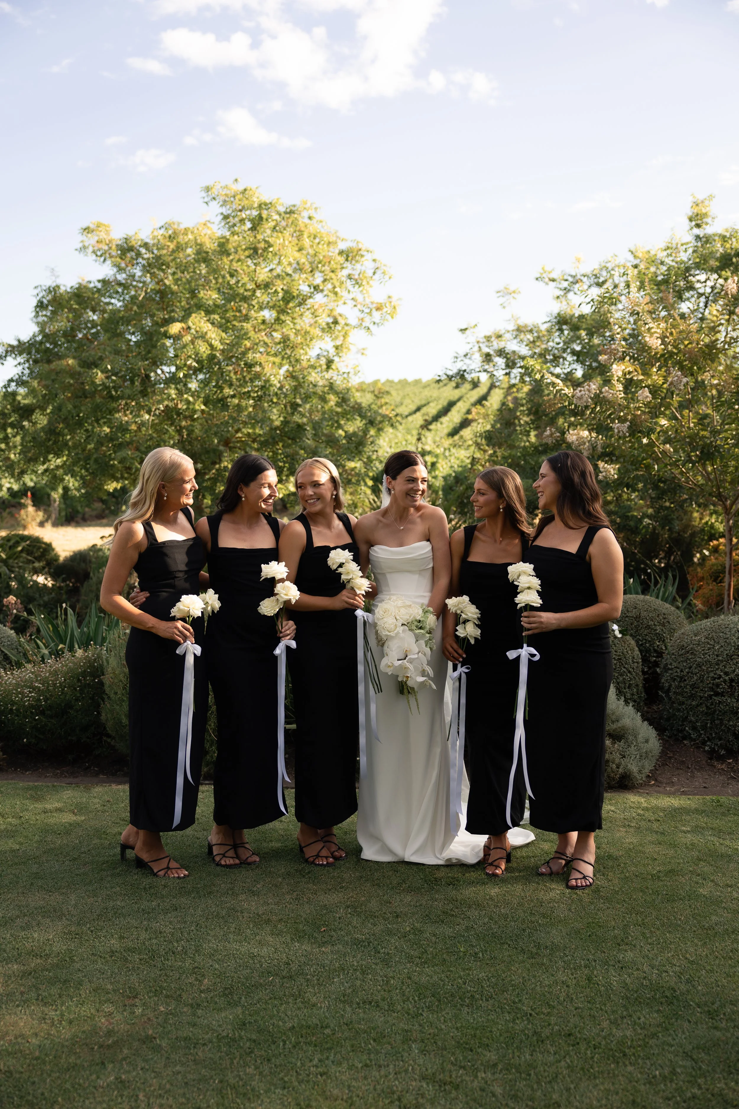A bride in a white dress standing with five bridesmaids in black dresses outdoors on a sunny day, all holding white flower bouquets, against a backdrop of green trees and shrubs.