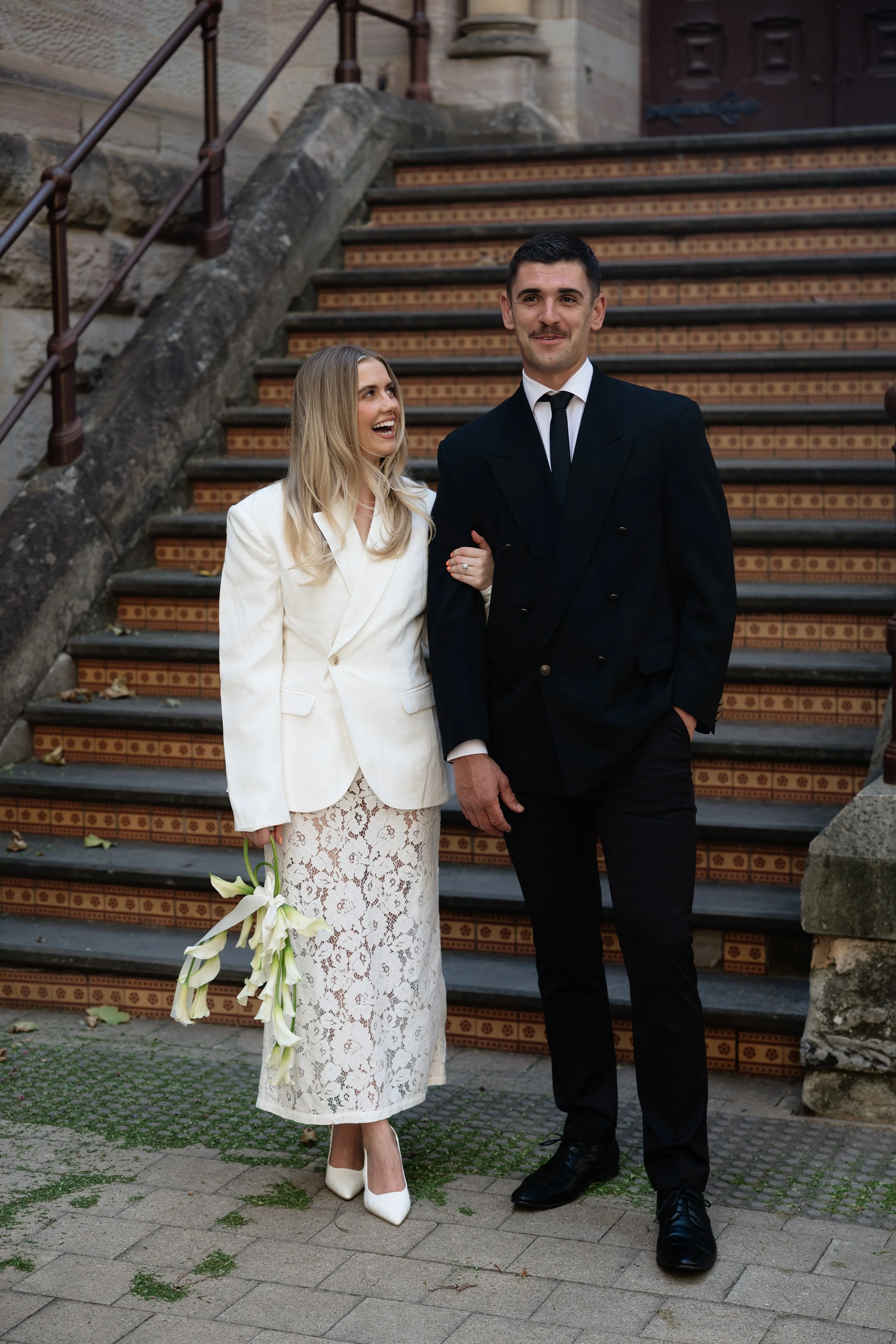 A smiling woman in a white blazer and lace skirt holding a bouquet of calla lilies, standing next to a man in a black suit and tie, on a staircase outside a stone building.