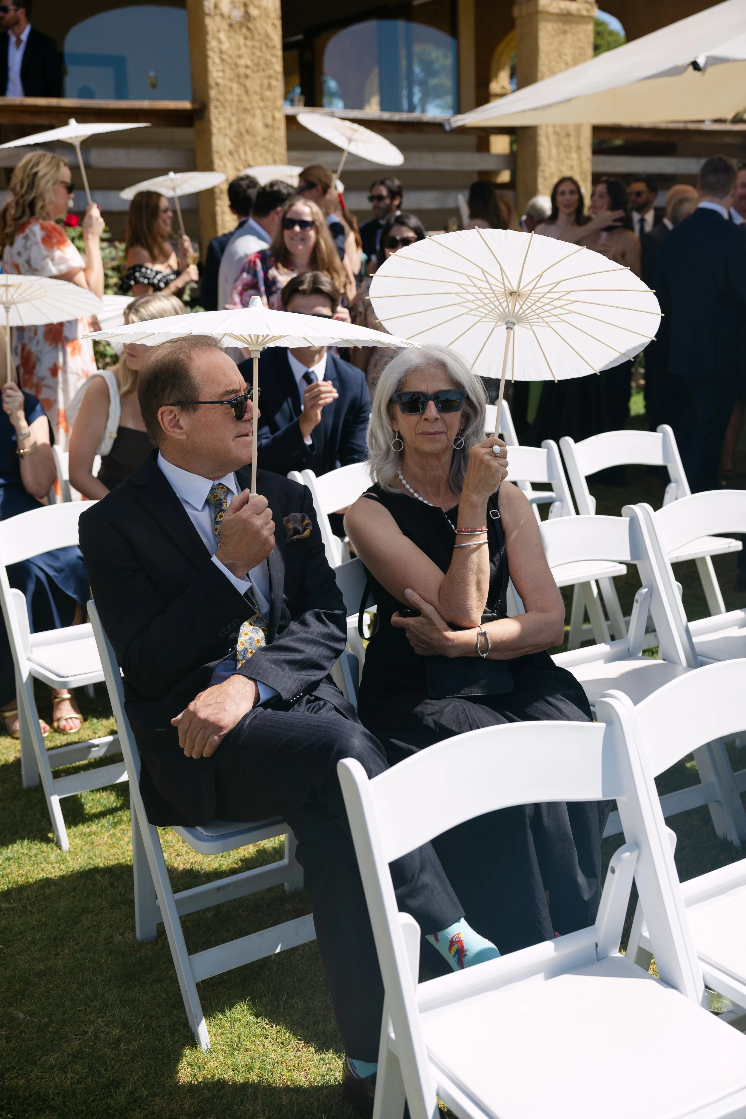 Two elderly people sitting outdoors at a formal event, holding white parasols, with other guests in the background.