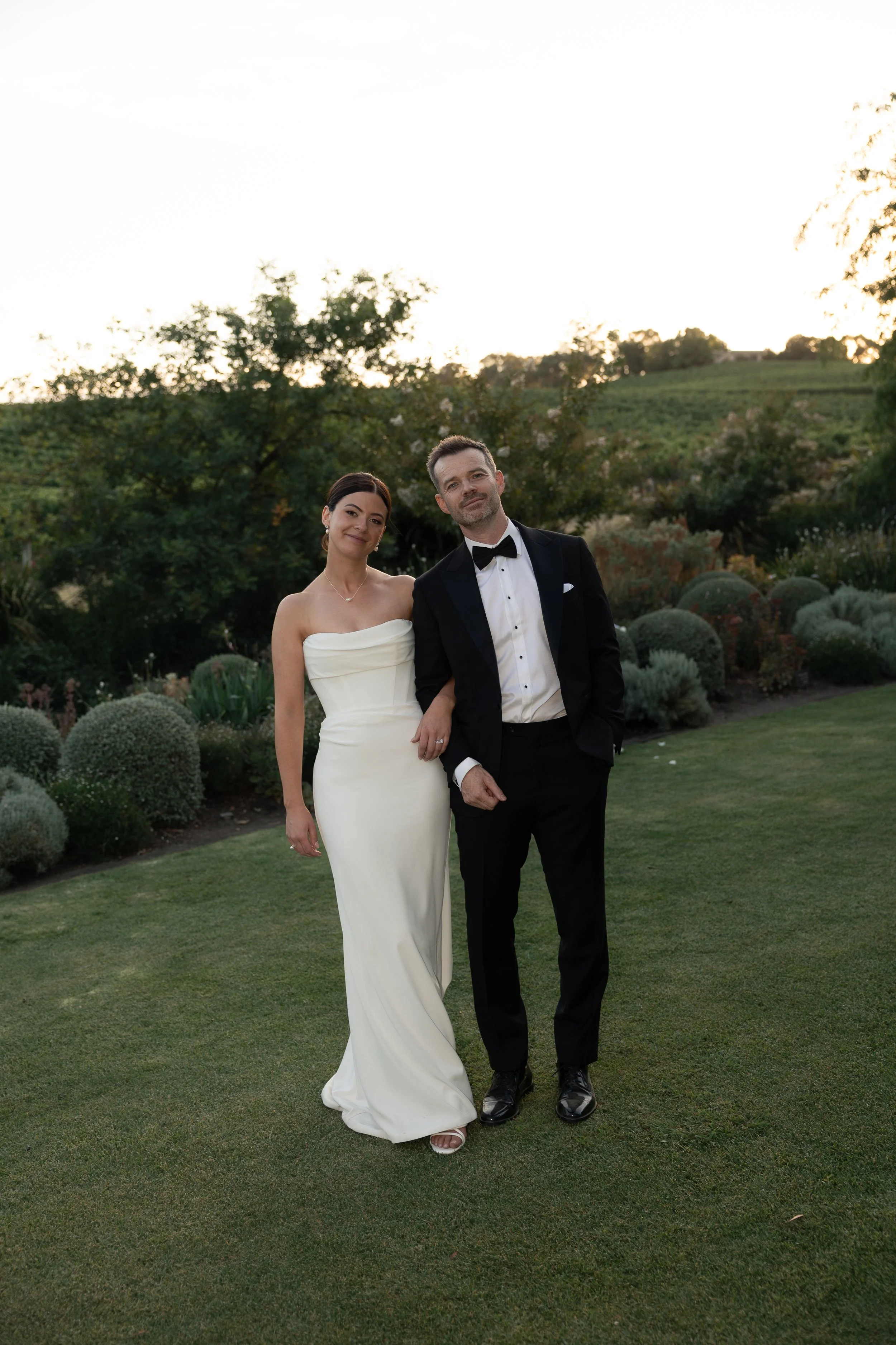 A bride and groom standing on a grass lawn outdoors during sunset, with the bride in a strapless white wedding gown and the groom in a black tuxedo with a bow tie, holding hands and smiling.