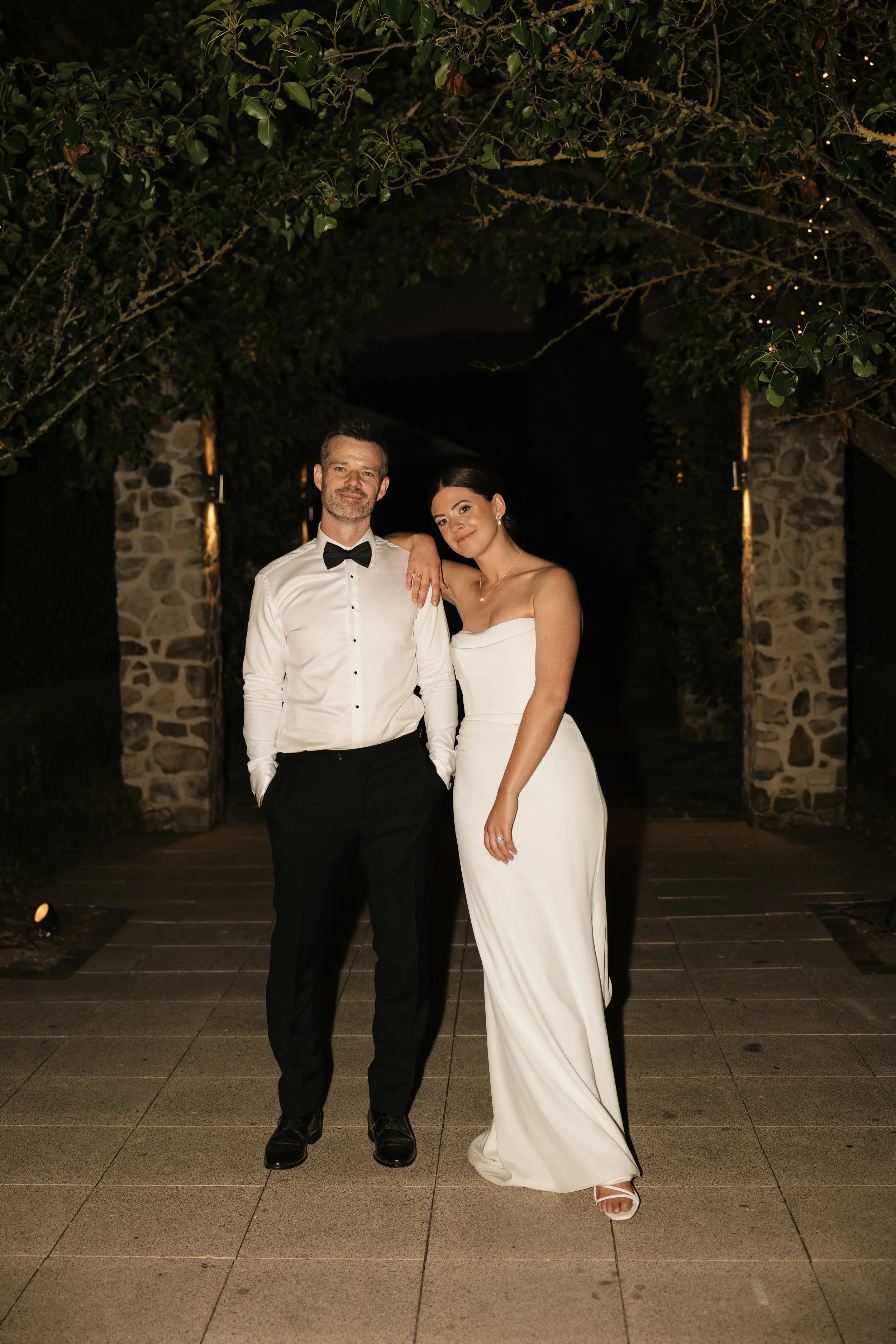 A man and woman in formal attire posing together at night outdoors, with the man in a black tuxedo and bowtie, and the woman in a strapless white gown, standing underneath a stone archway with greenery.