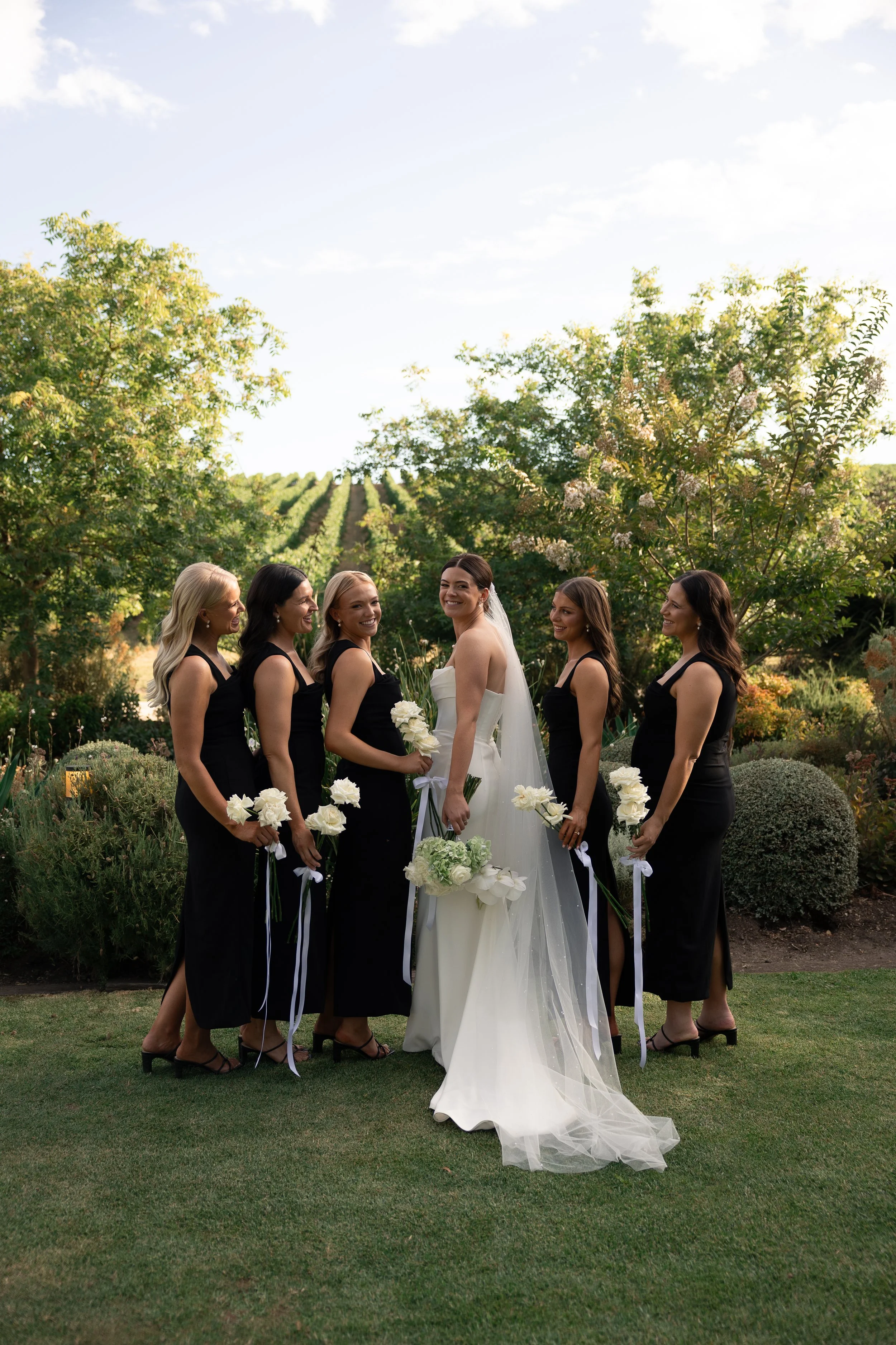 A bride and five bridesmaids standing outdoors in a garden with lush greenery, trees, and rolling hills in the background. The bride, in a white wedding gown, is holding a bouquet, while the bridesmaids are in black dresses, each holding a white flow