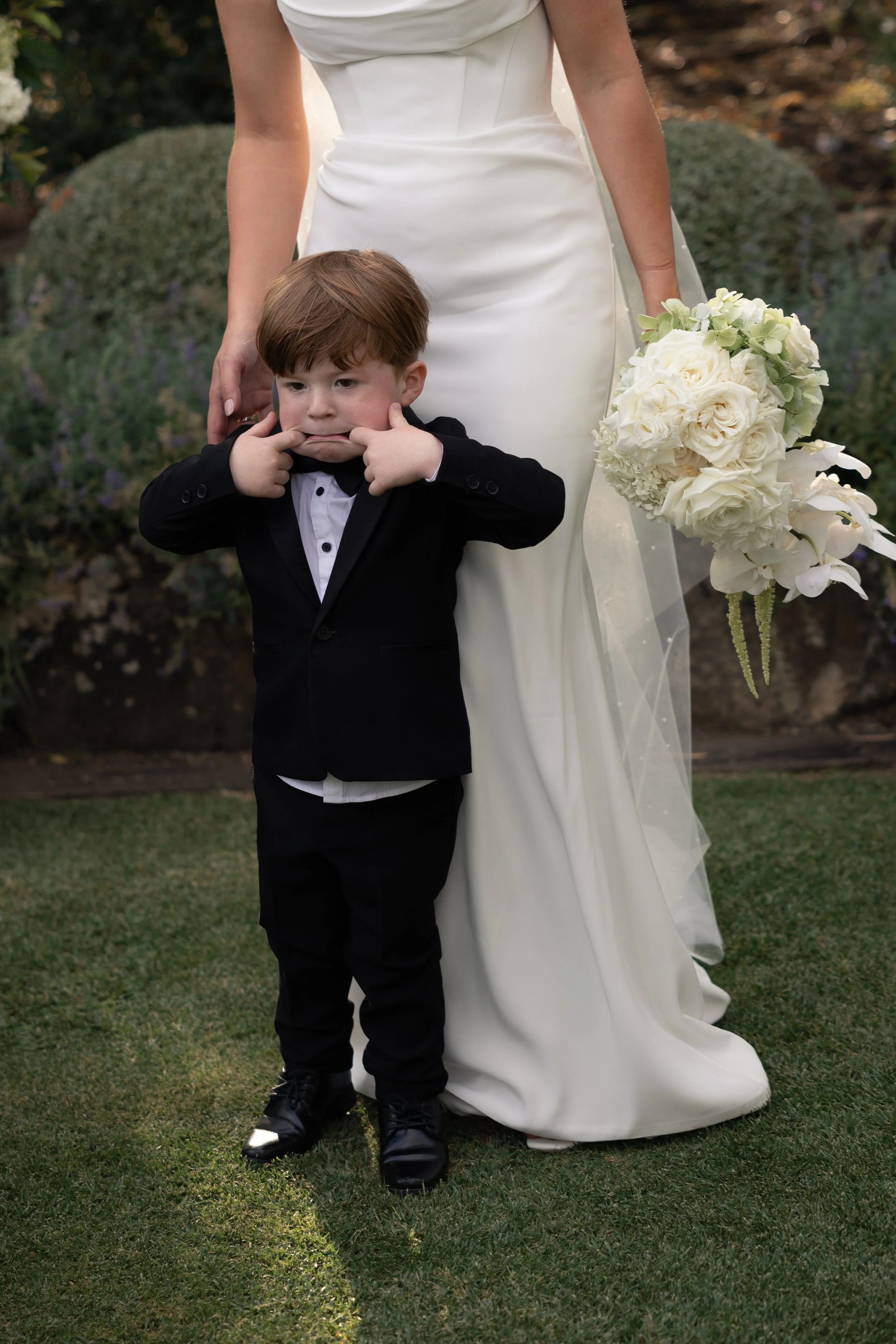 A young boy in a tuxedo making a face with his fingers in his mouth, standing next to a woman in a white wedding dress holding a bouquet of white flowers.