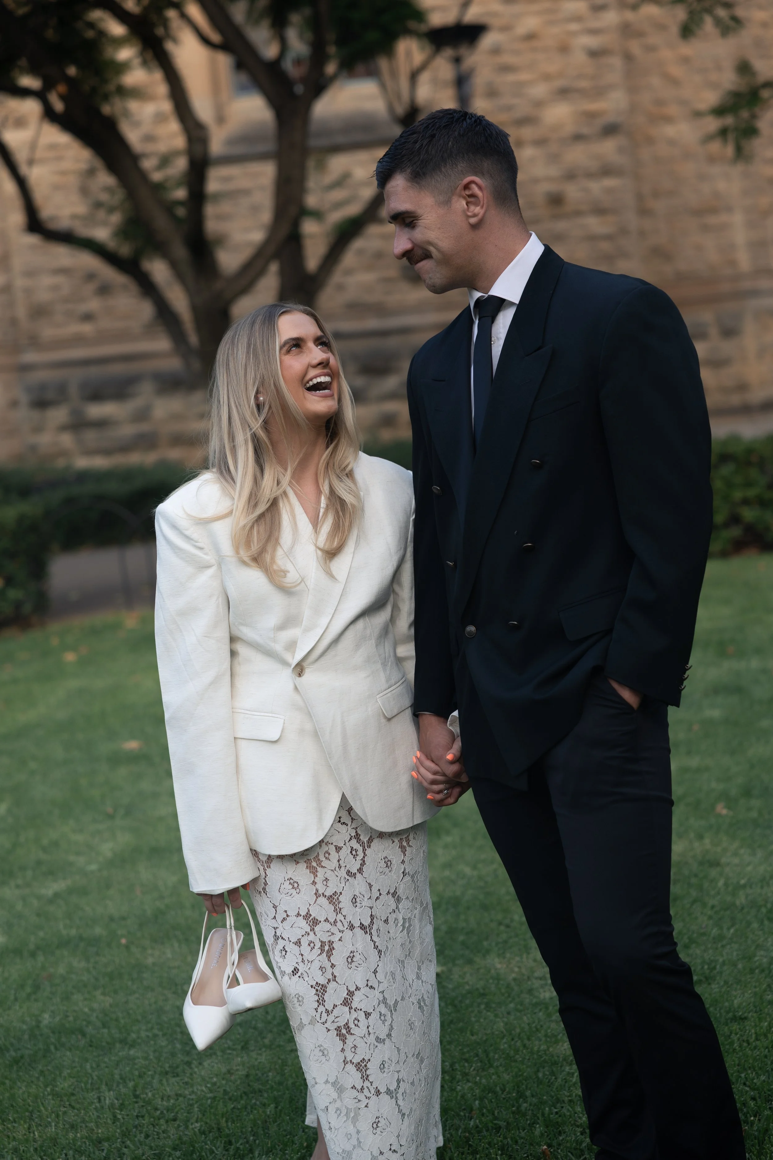 A woman in a white blazer and lace skirt holding white high-heeled shoes, smiling and looking up at a man in a black suit, who is holding her hand in an outdoor setting with grass and trees.