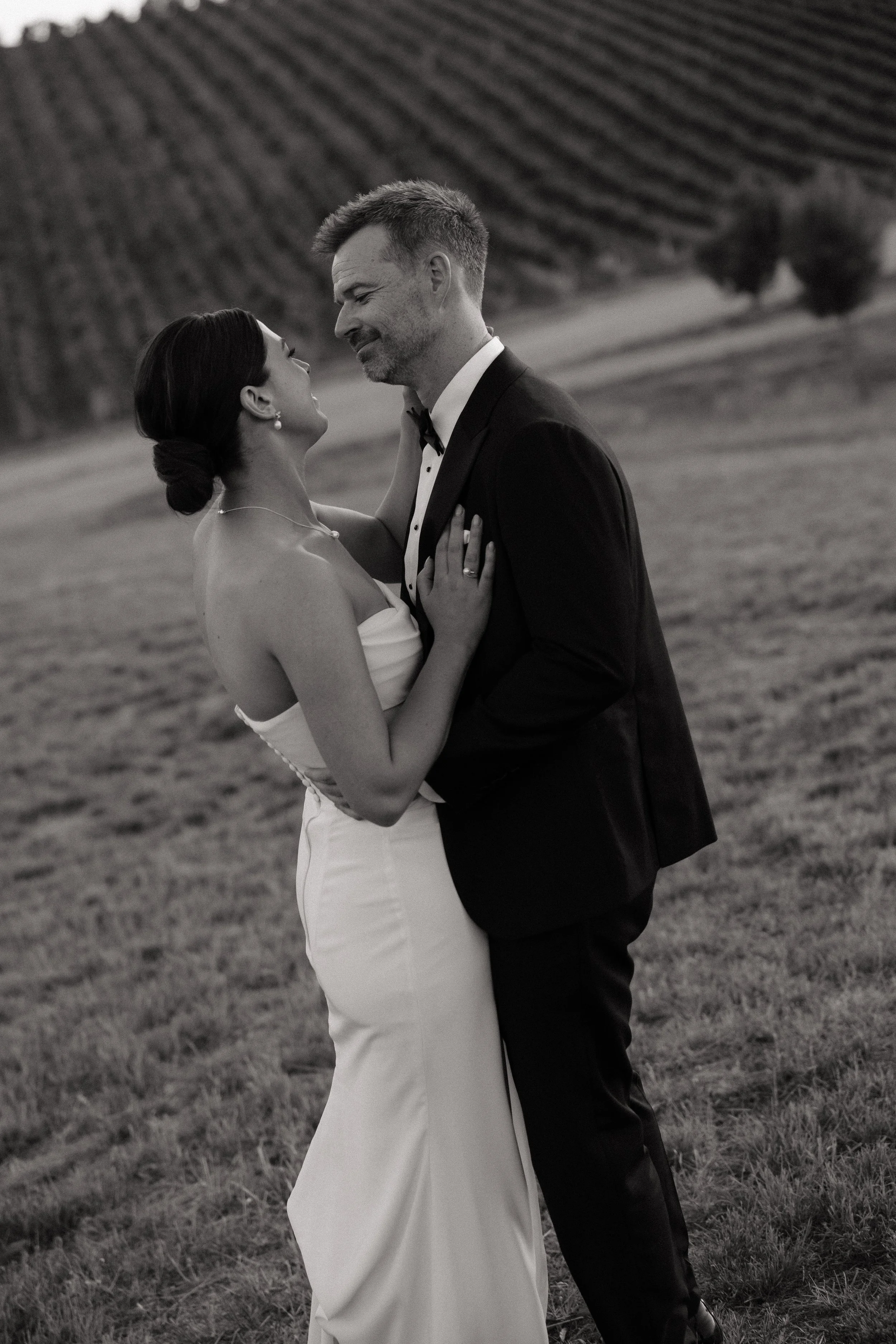 A black-and-white photo of a bride and groom embracing outdoors, with a field and rolling hills in the background.