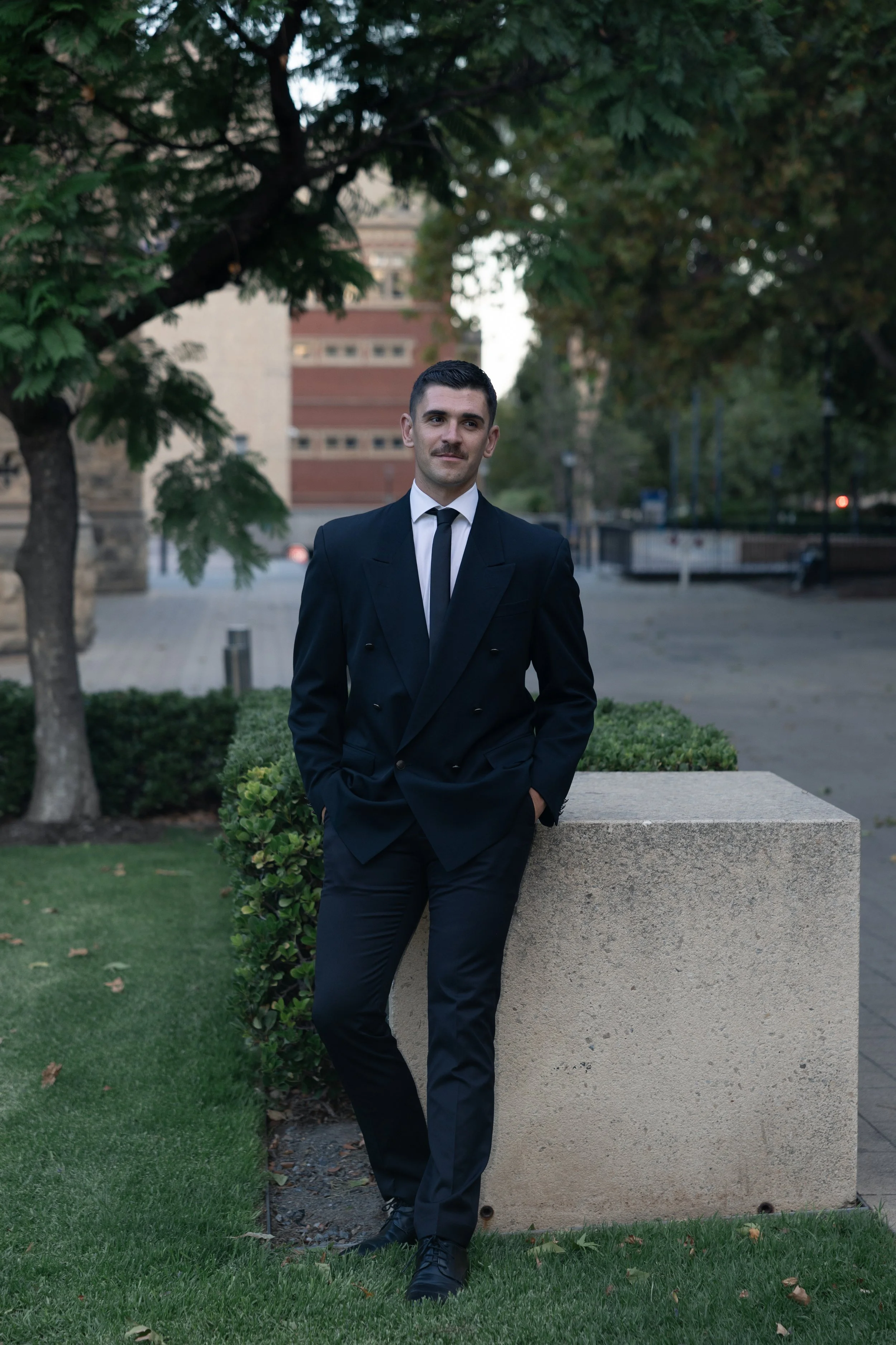A man in a black suit, white shirt, and black tie standing outdoors next to a concrete block, with trees and buildings in the background.
