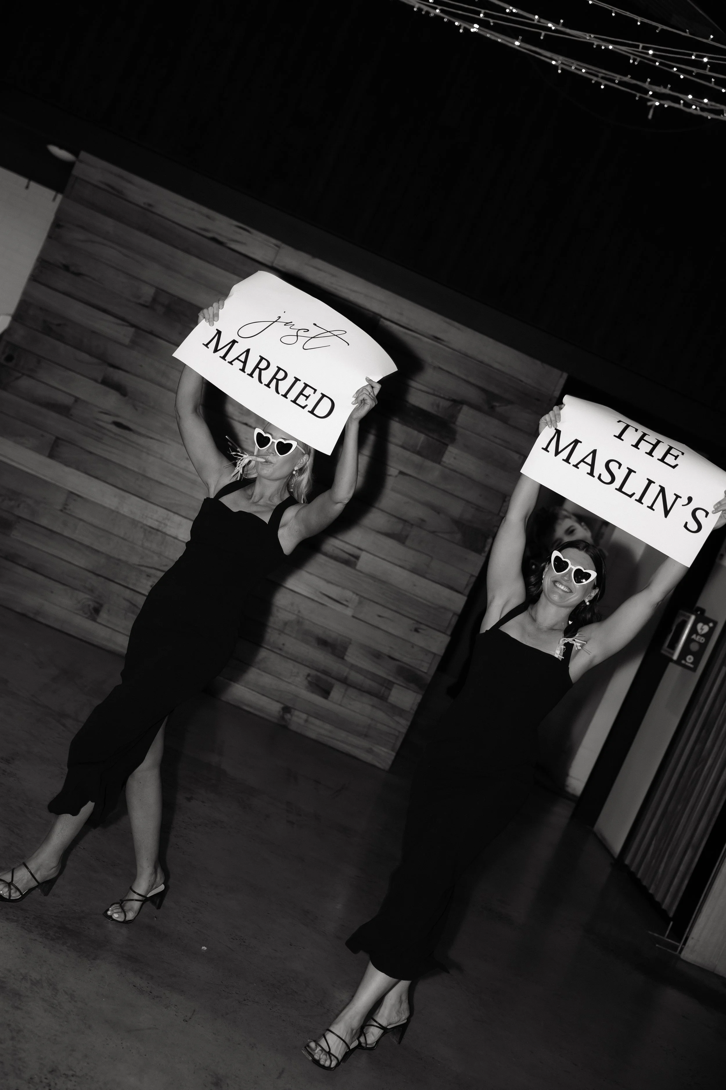 Two women in black dresses and high heels holding signs that say 'Just MARRIED' and 'THE MASCULIN'S'. They are wearing large heart-shaped sunglasses and celebrating a wedding indoors with wooden panel walls.