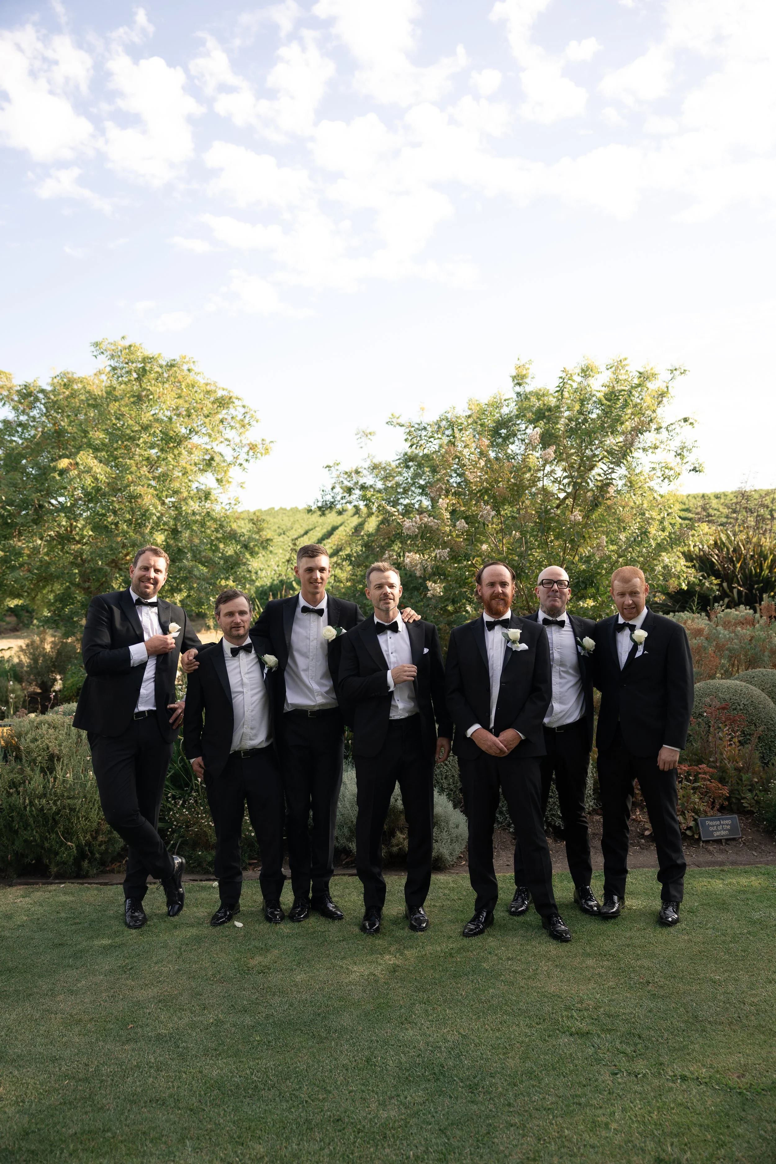 A group of seven men dressed in black tuxedos with white shirts and black bow ties, standing outdoors in a garden, posing for a photo, some with white roses on their lapels, under a partly cloudy sky.