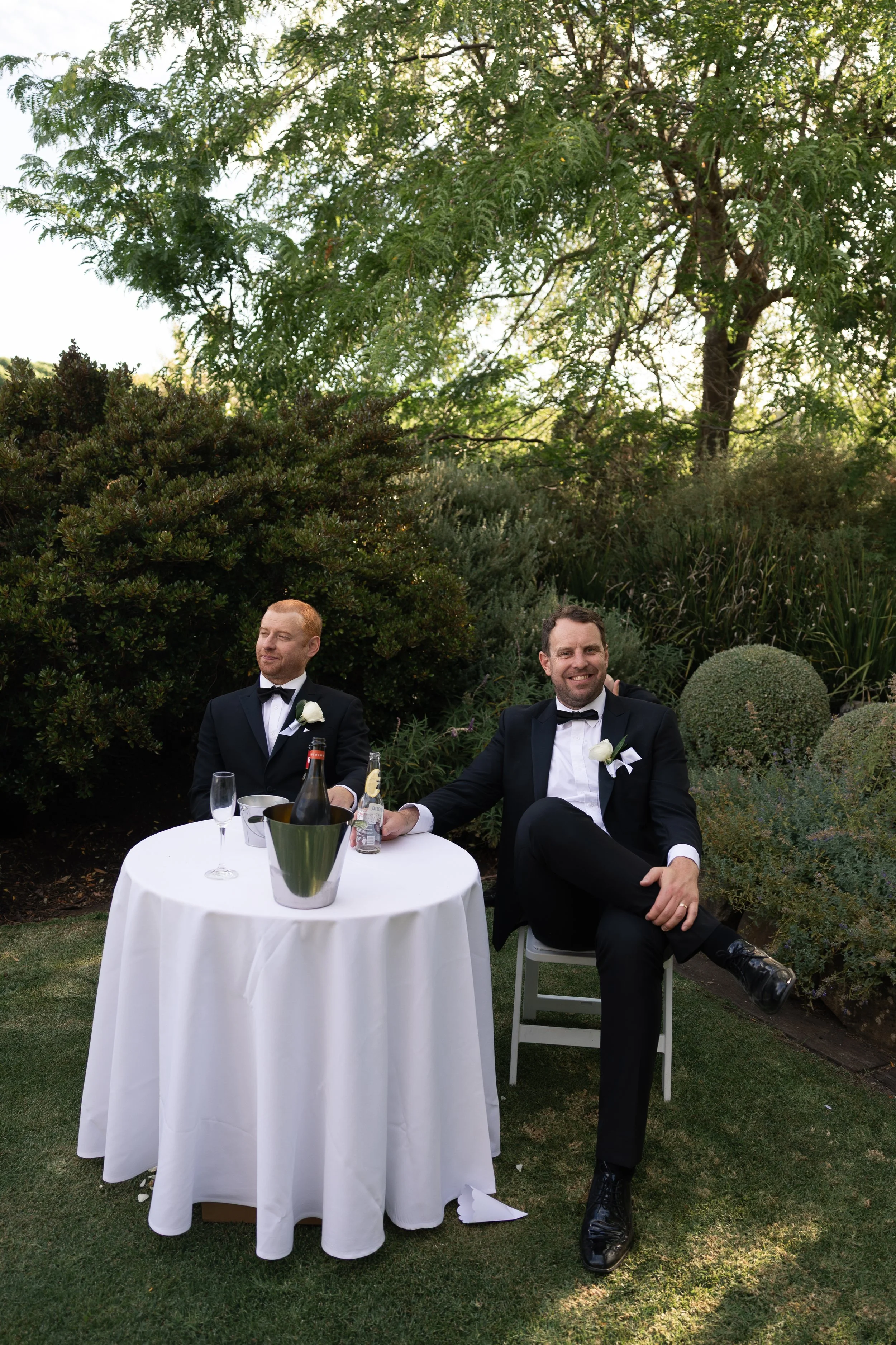 Two men in tuxedos sitting at a round table with a white tablecloth outdoors, smiling. The table has a champagne bottle, glasses, and a bucket. One man is smiling directly at the camera, the other is looking away. Lush green trees and bushes surround