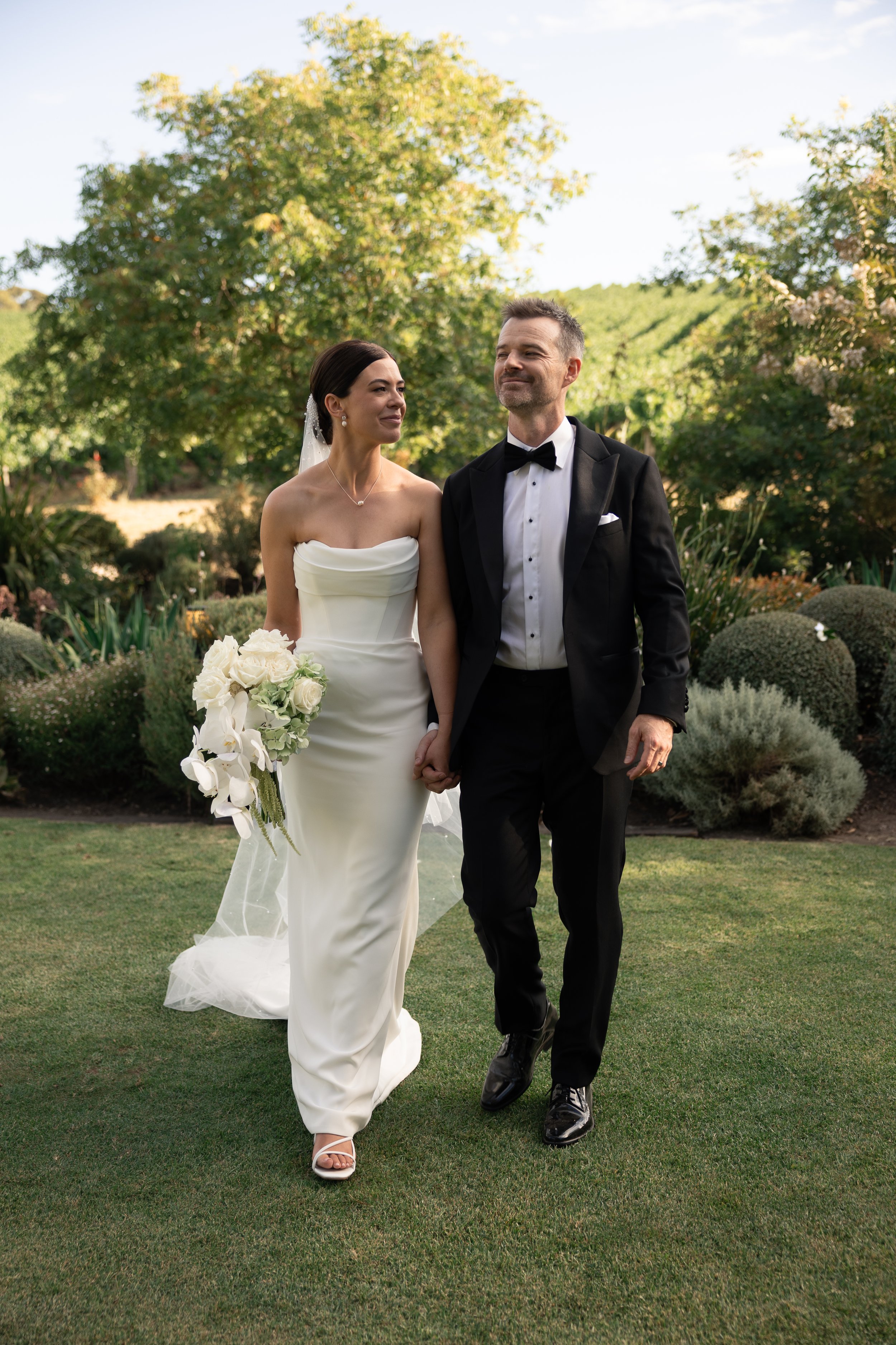 A newlywed couple walking hand-in-hand on grass in a garden, with the bride in a strapless white wedding gown holding a bouquet and the groom in a black tuxedo with a bow tie, both smiling and looking content.