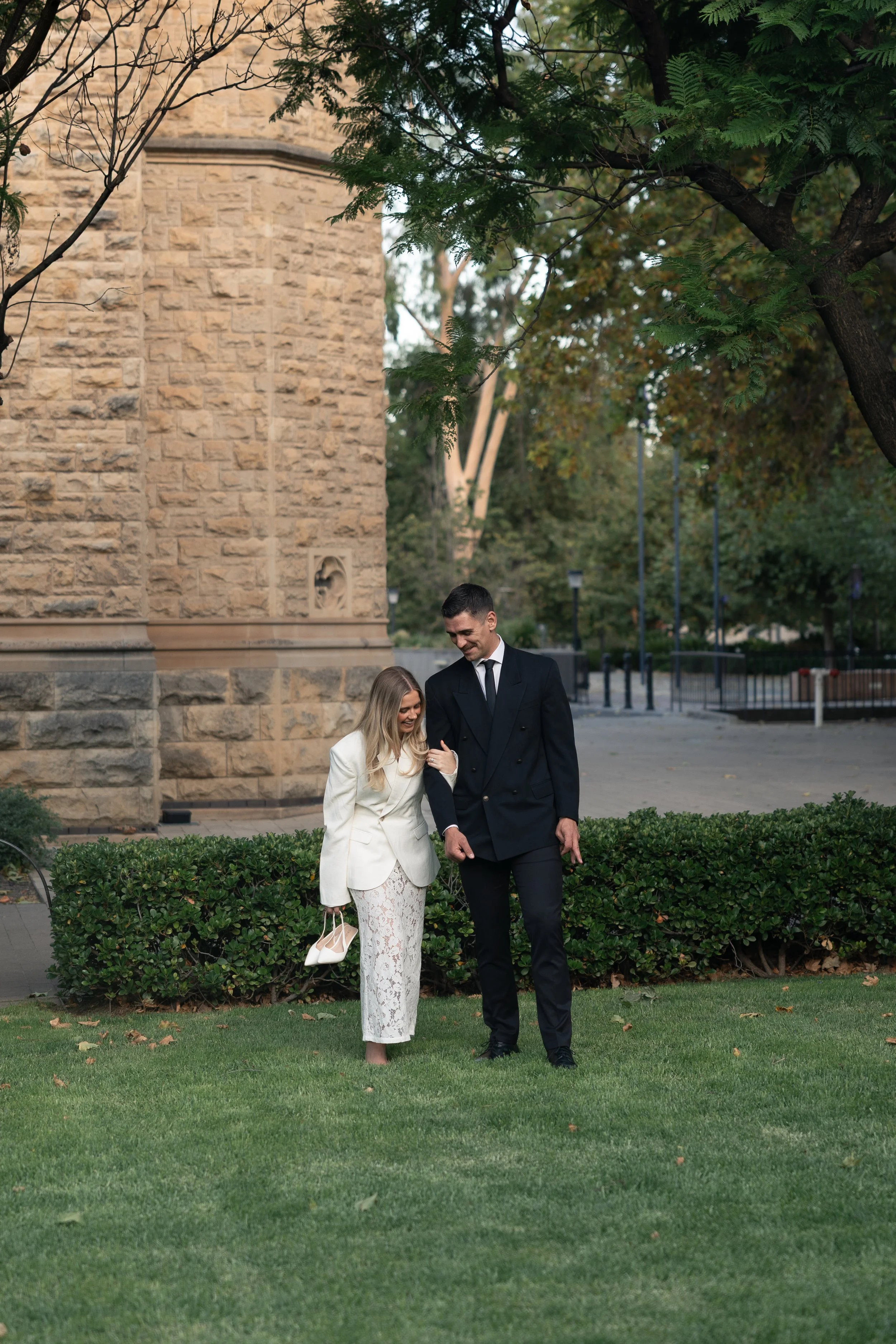 A woman in white pants and a white blazer holding white shoes walking with a man in a black suit on a grassy area in front of a stone building during daytime.