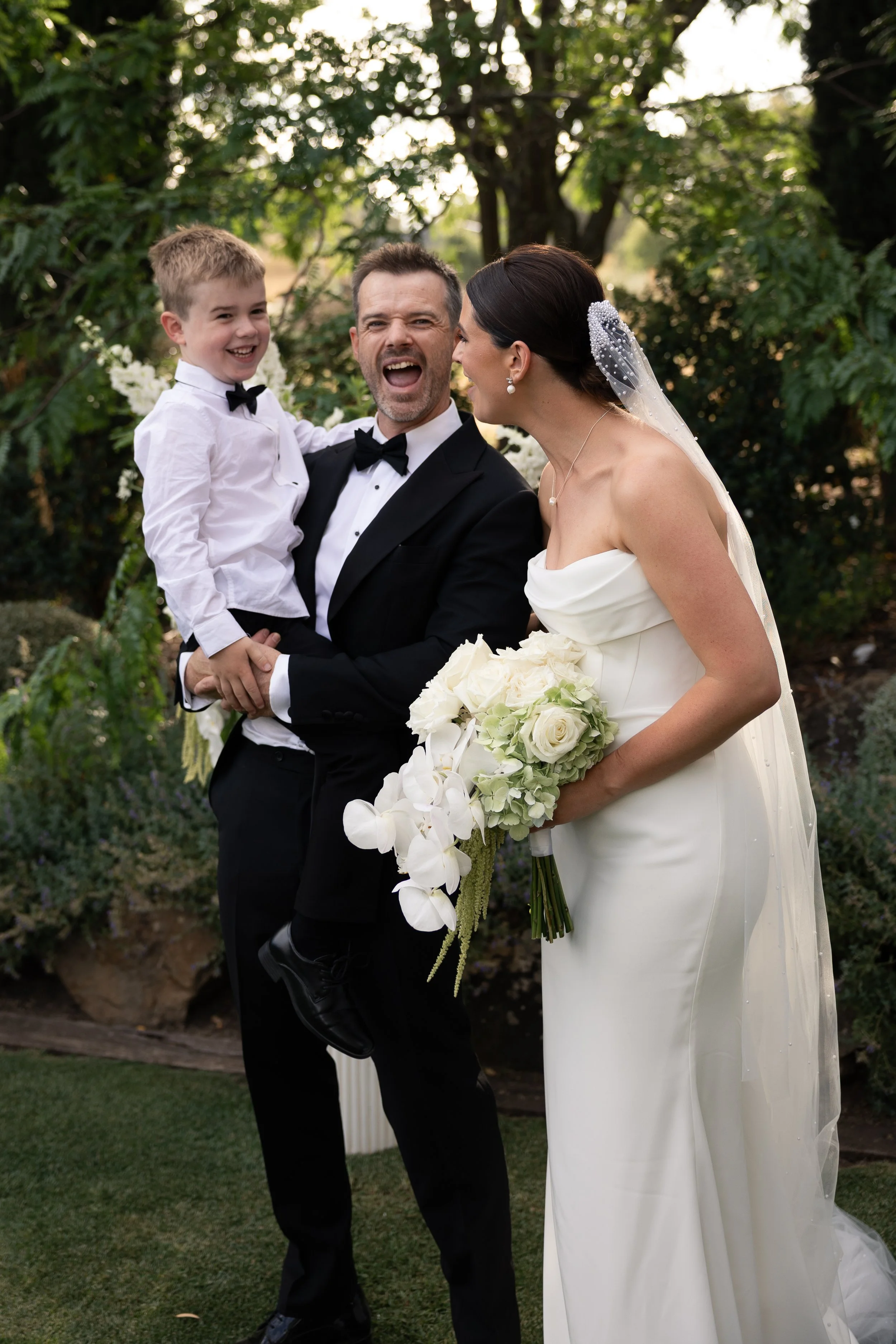 A wedding scene with a bride and groom, the groom holding a young boy, all smiling and appearing happy, outdoors with greenery in the background.