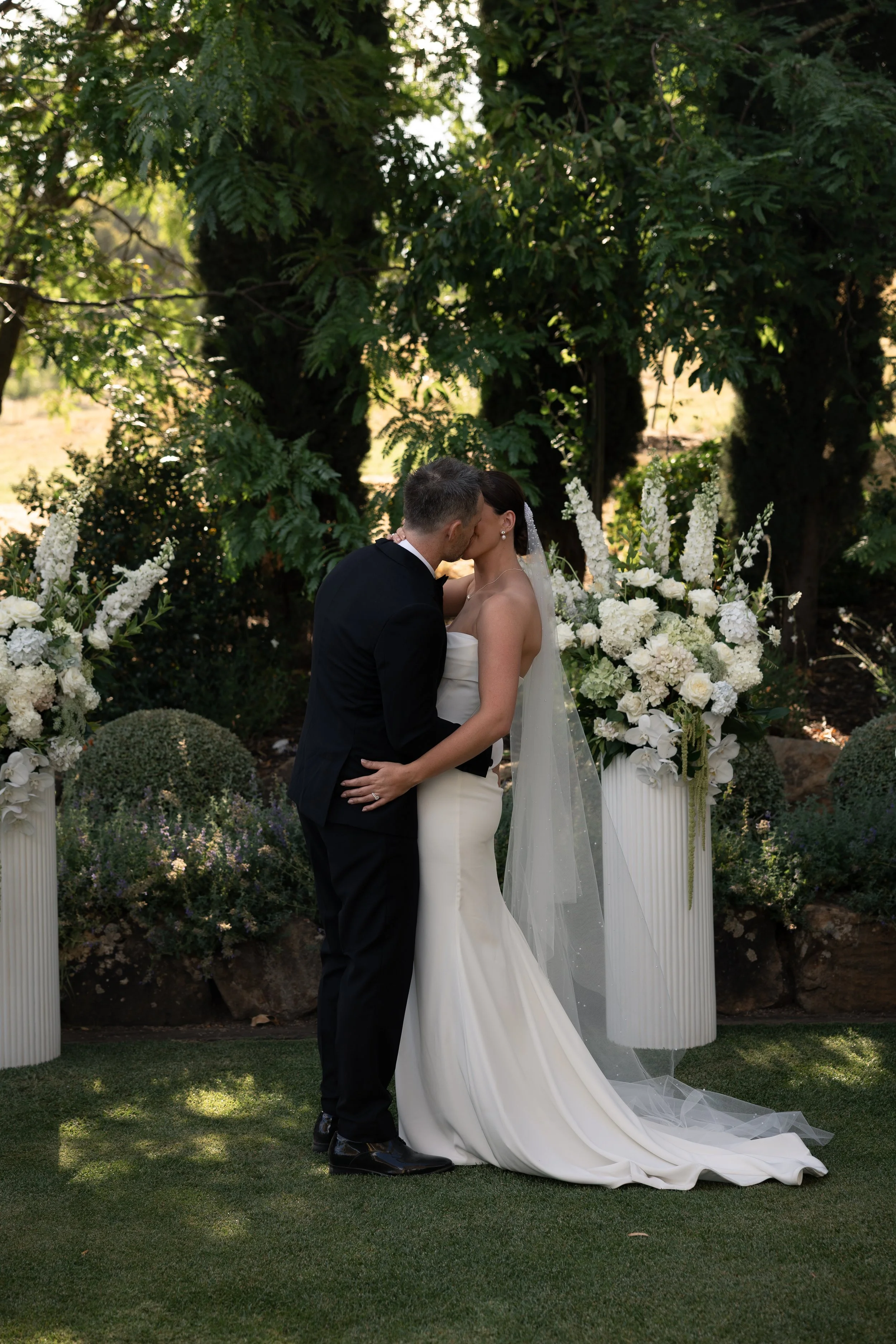 A bride and groom share a kiss in an outdoor wedding ceremony surrounded by white floral arrangements and lush greenery.
