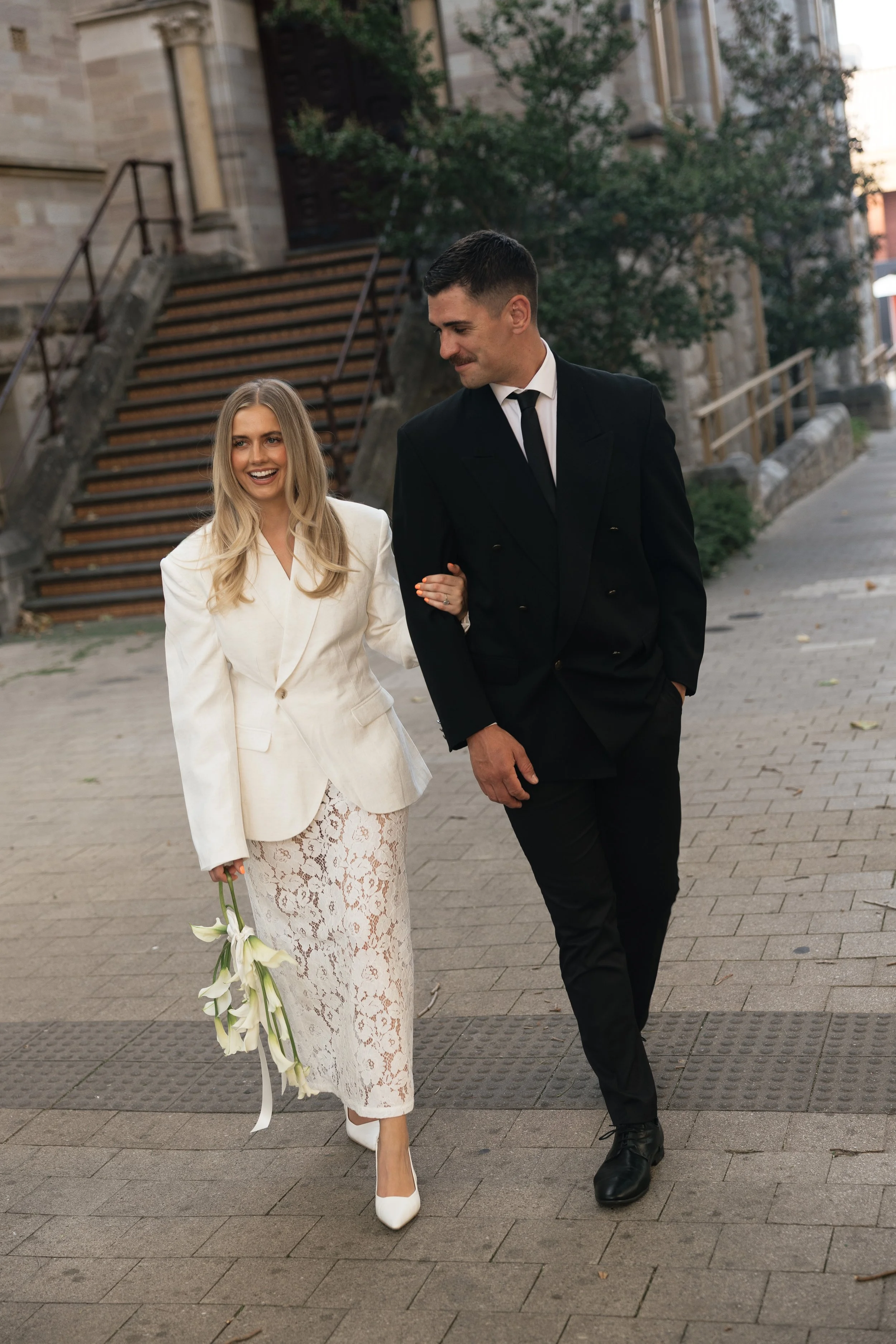A man and woman walking arm-in-arm on a city sidewalk, dressed in formal attire; the woman is holding white calla lilies and wearing a white lace dress and blazer, the man is in a black suit and tie.