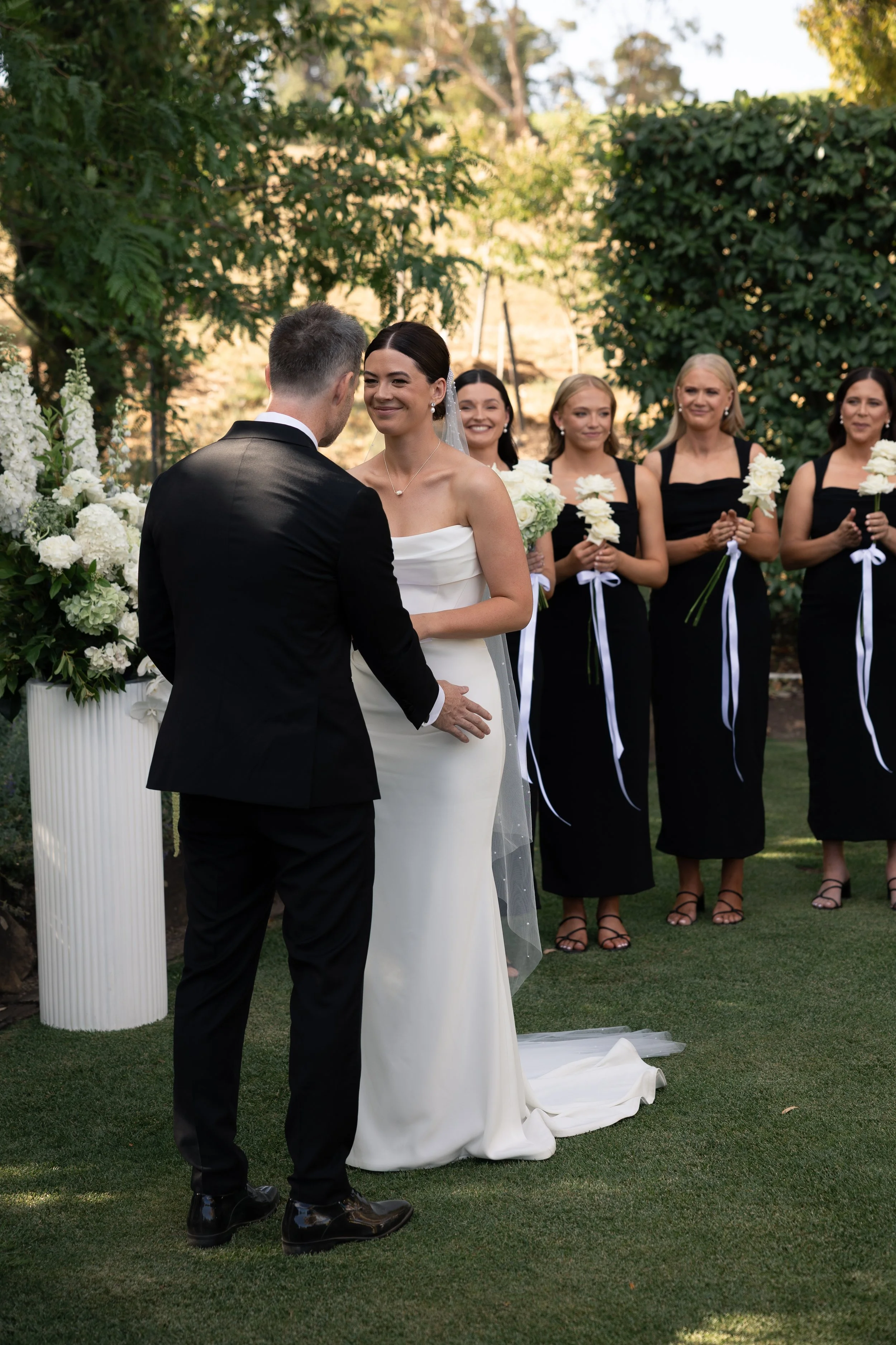 Bride and groom exchanging vows during outdoor wedding ceremony, with bridesmaids holding white flowers in the background.