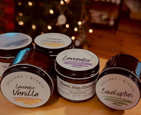 Collection of small black jars with labels including lavender vanilla, lavender lemongrass, eucalyptus, and lavender tallow moisturizer, arranged on a wooden surface with Christmas tree in the blurred background.