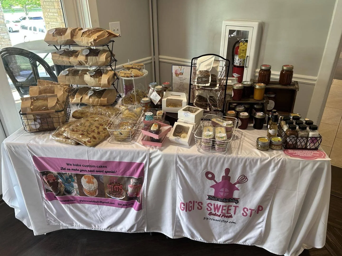 A table with various baked goods, jars of jam, and baked treats from Gigi's Sweet Stop, set up in a corner near a window and fire safety equipment.