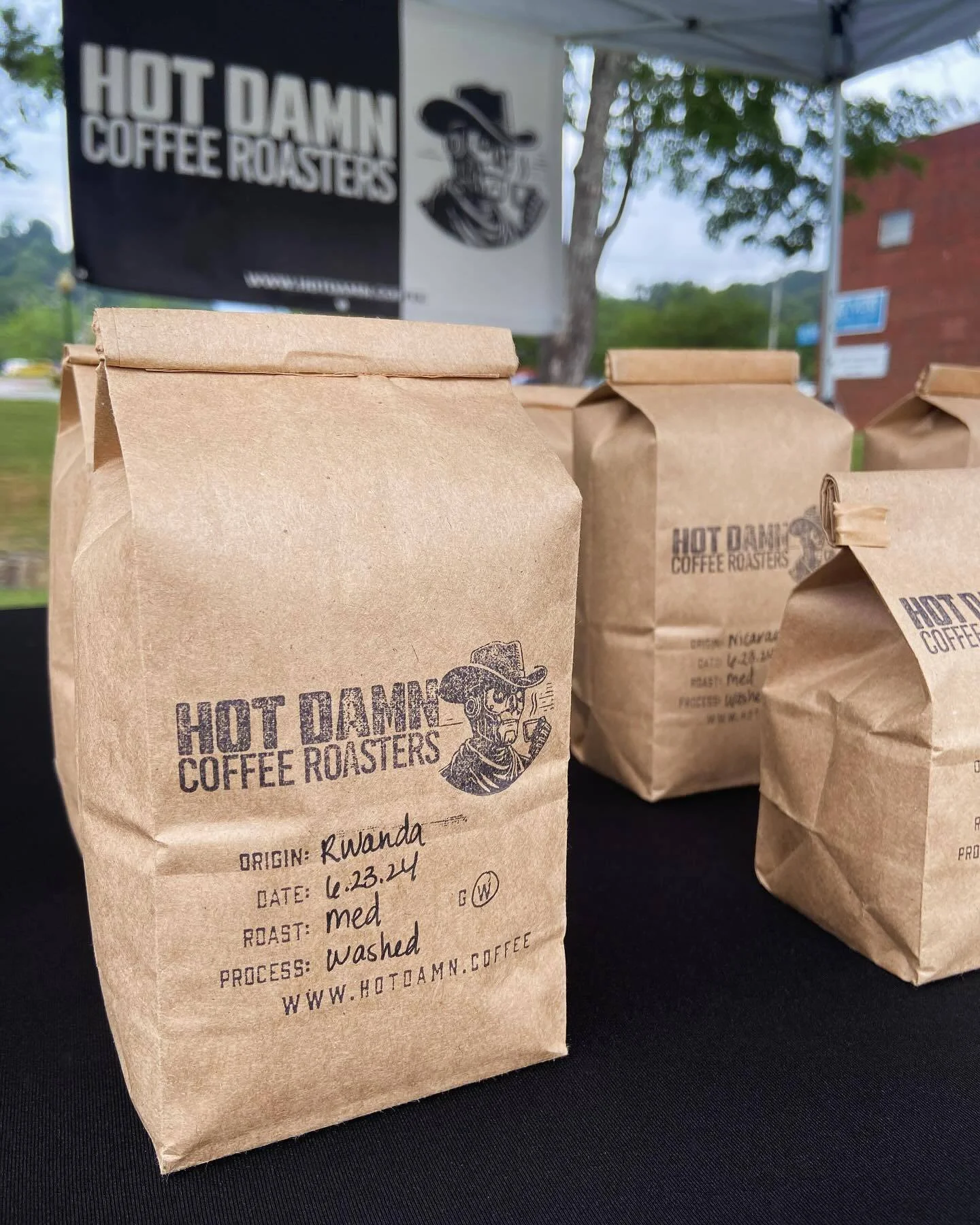 Several brown paper bags of coffee labeled with origin, date, roast level, and process, in front of a banner for Hot Damn Coffee Roasters, outdoors under a white canopy.