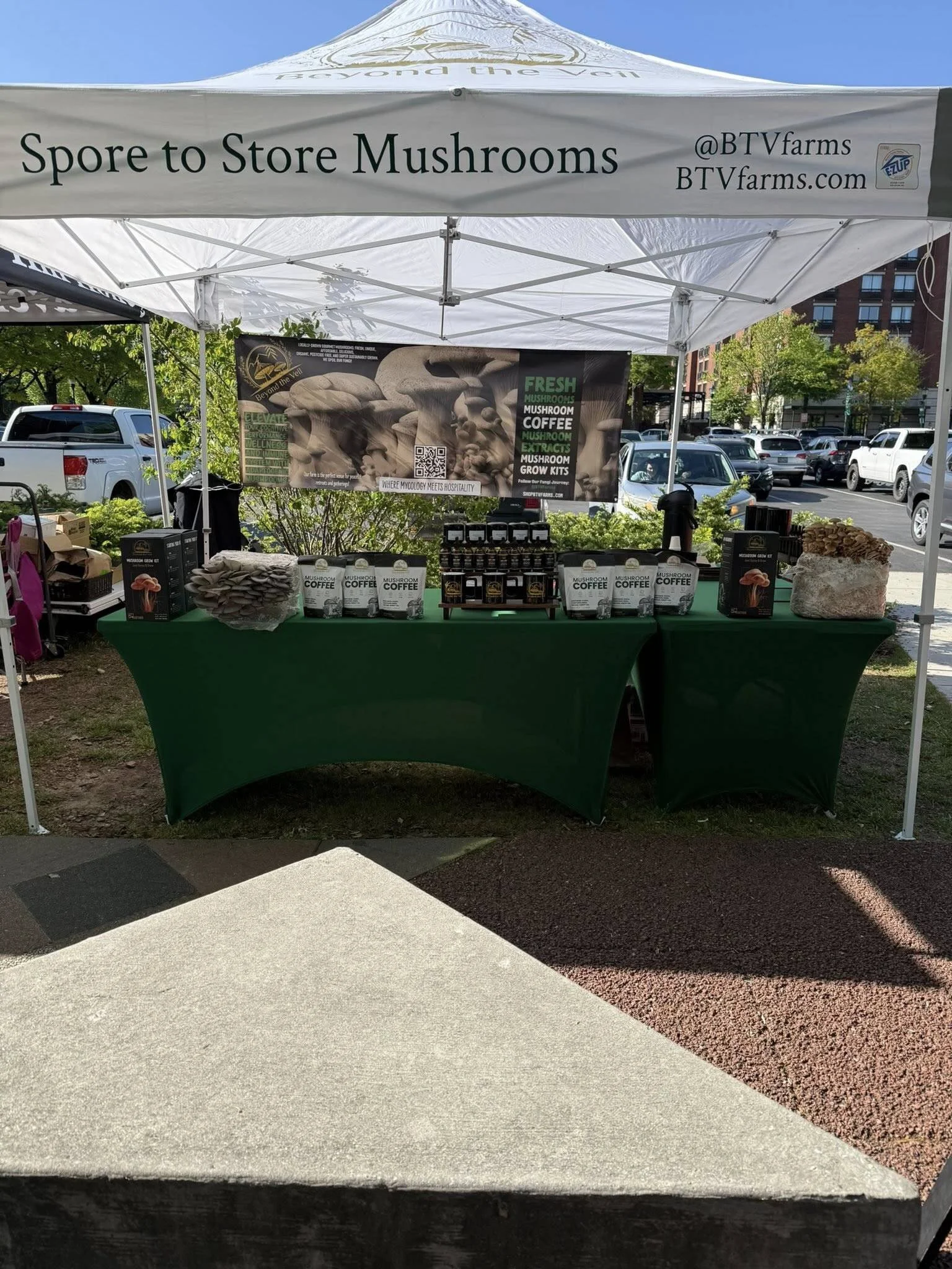 Market tent with a sign that reads 'Spore to Store Mushrooms' and offers fresh mushrooms, mushroom coffee, extracts, and grow kits, with plants and parked cars in the background.