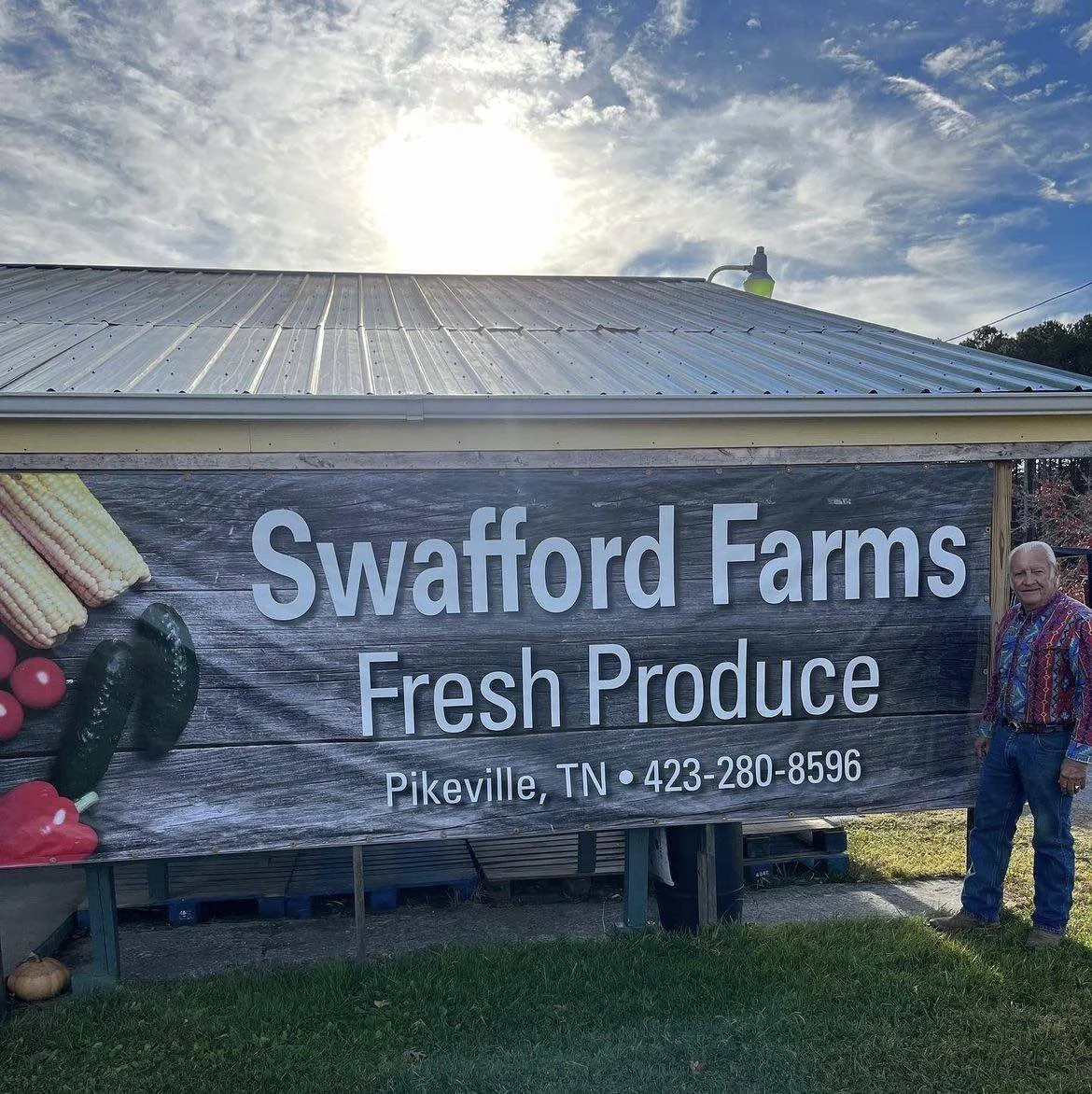 Sign for Swafford Farms, a fresh produce market in Pikeville, Tennessee. The sign features images of corn, peppers, and tomatoes, with a man standing next to it during daytime with a partly cloudy sky.