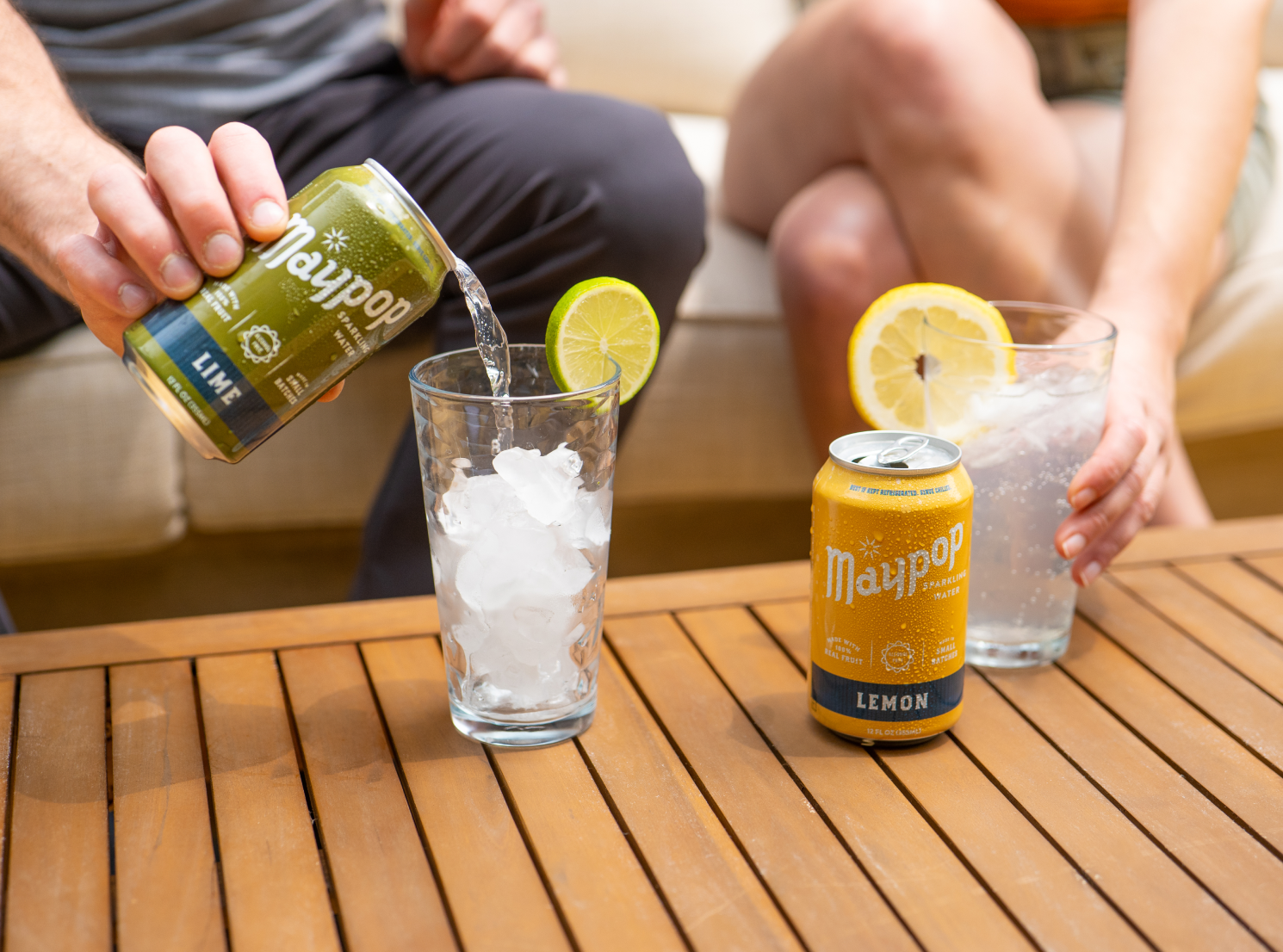 Person pouring lime-flavored sparkling water from a Maypop can into a glass with ice and a lime wedge, while another person holds a glass with a lemon wedge on a wooden table.