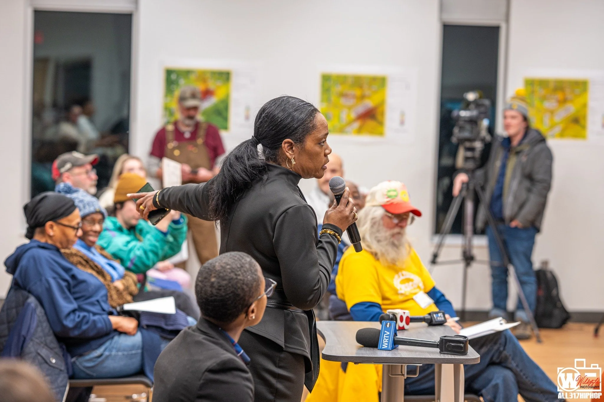 A woman speaking into a microphone during a community event or meeting, with a group of seated people and a cameraman recording in the background.