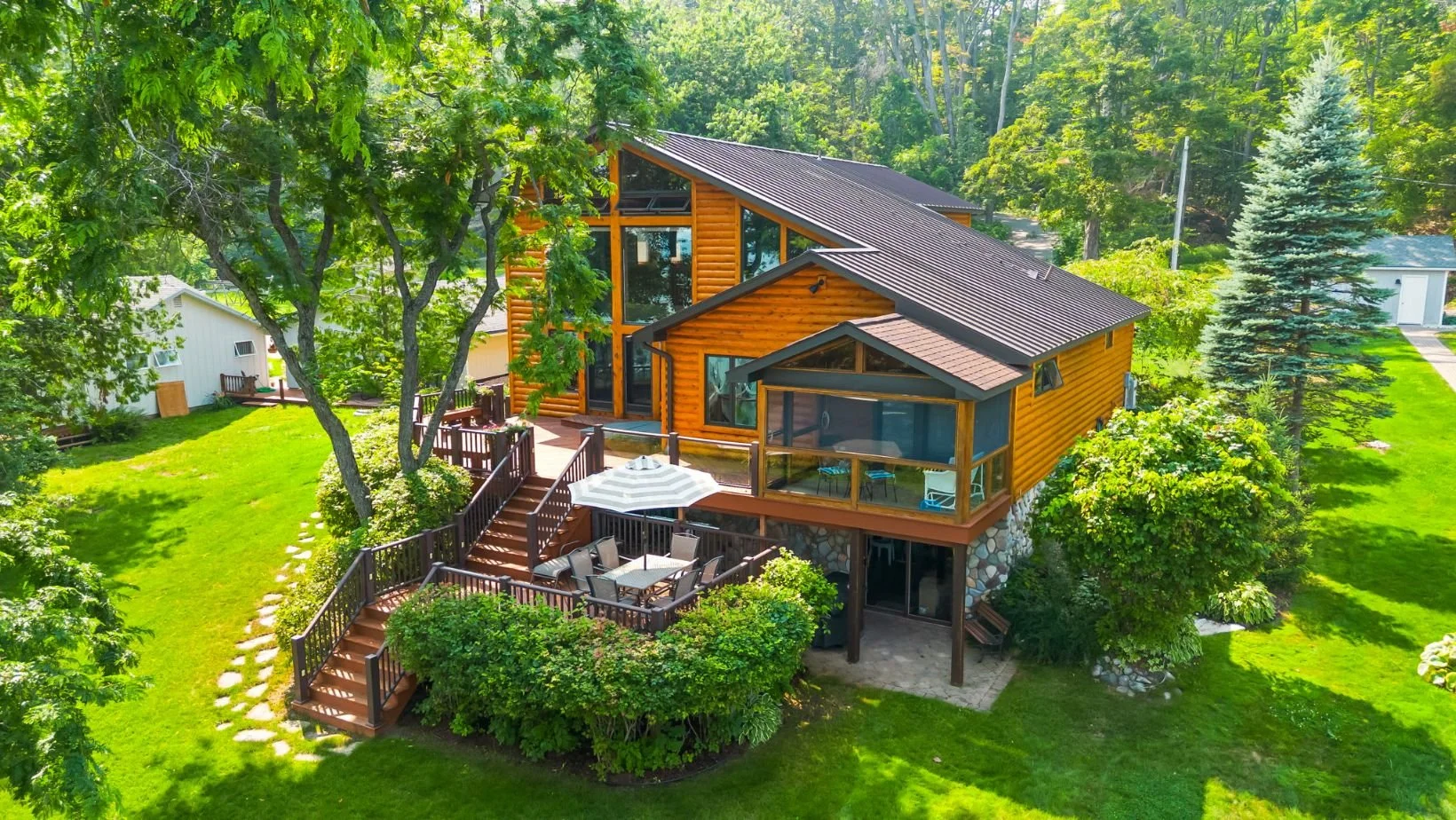 A modern, multi-level house with wooden siding and large windows, surrounded by green trees and a well-maintained lawn with a deck and outdoor dining area.