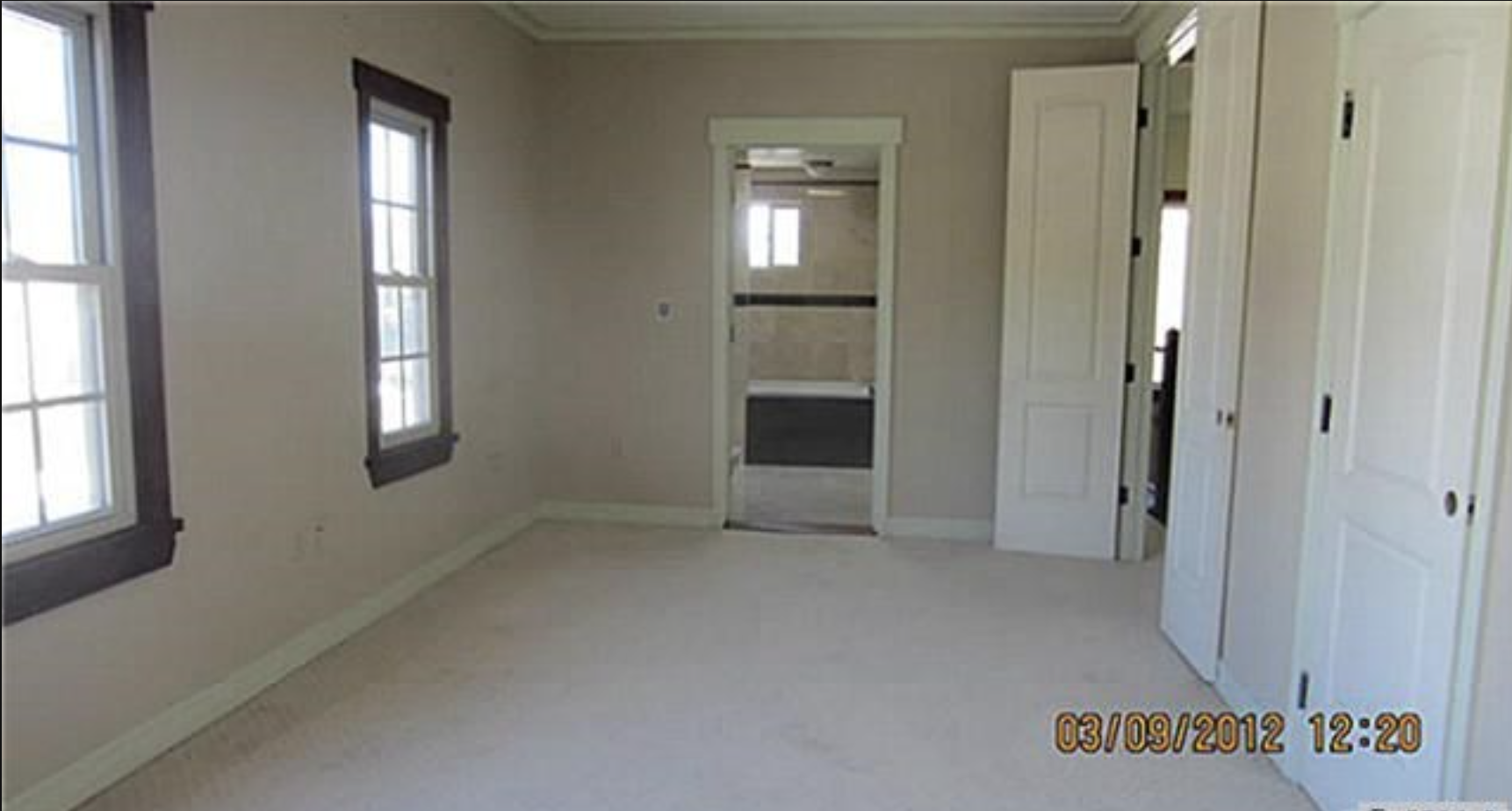 Empty room with three windows, beige walls, white trim, and double doors, with a view into a bathroom with a window, tiled walls, and a black accent stripe, and a closet with white doors.