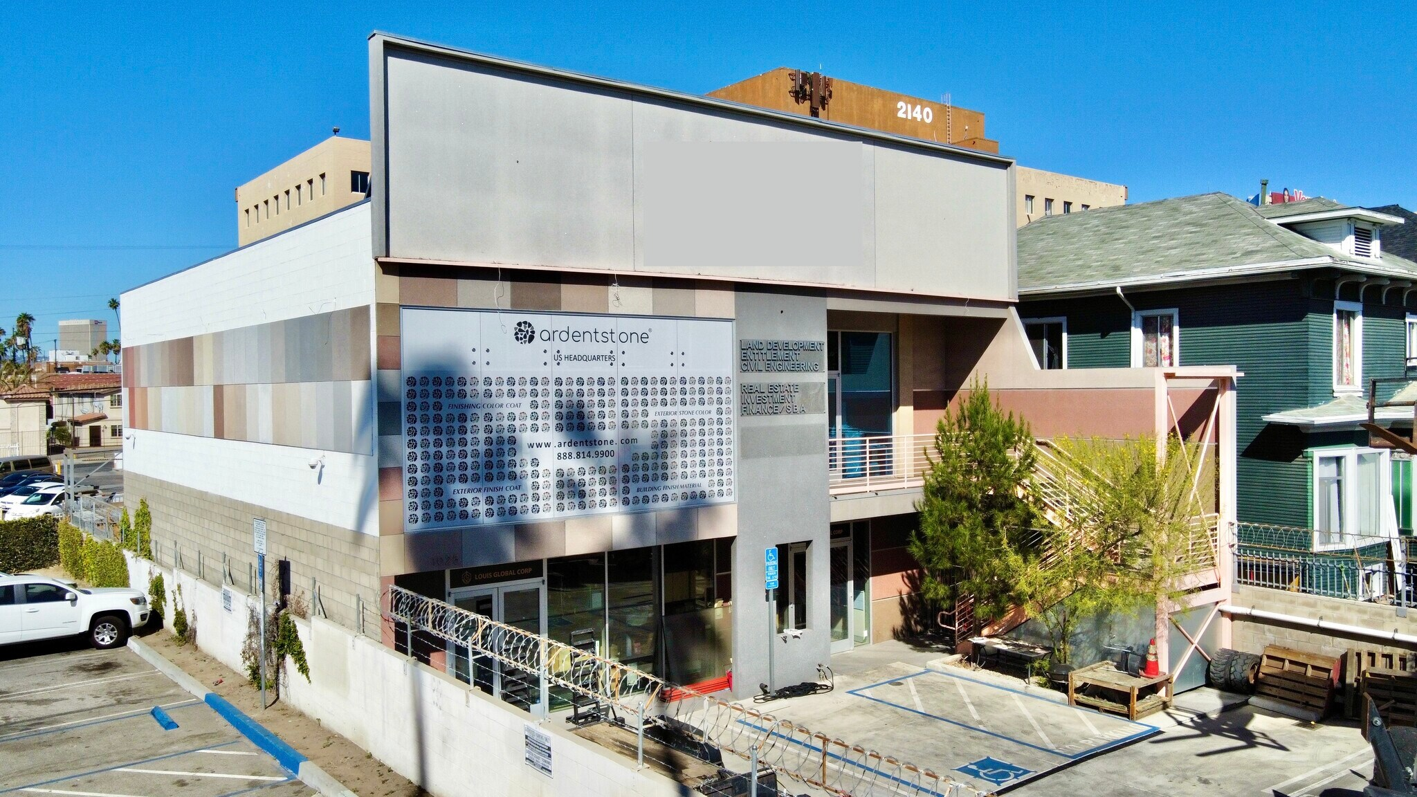Commercial building with sign for Ardent Stone in an urban area, with parking lot, trees, and neighboring houses visible, under a clear blue sky.