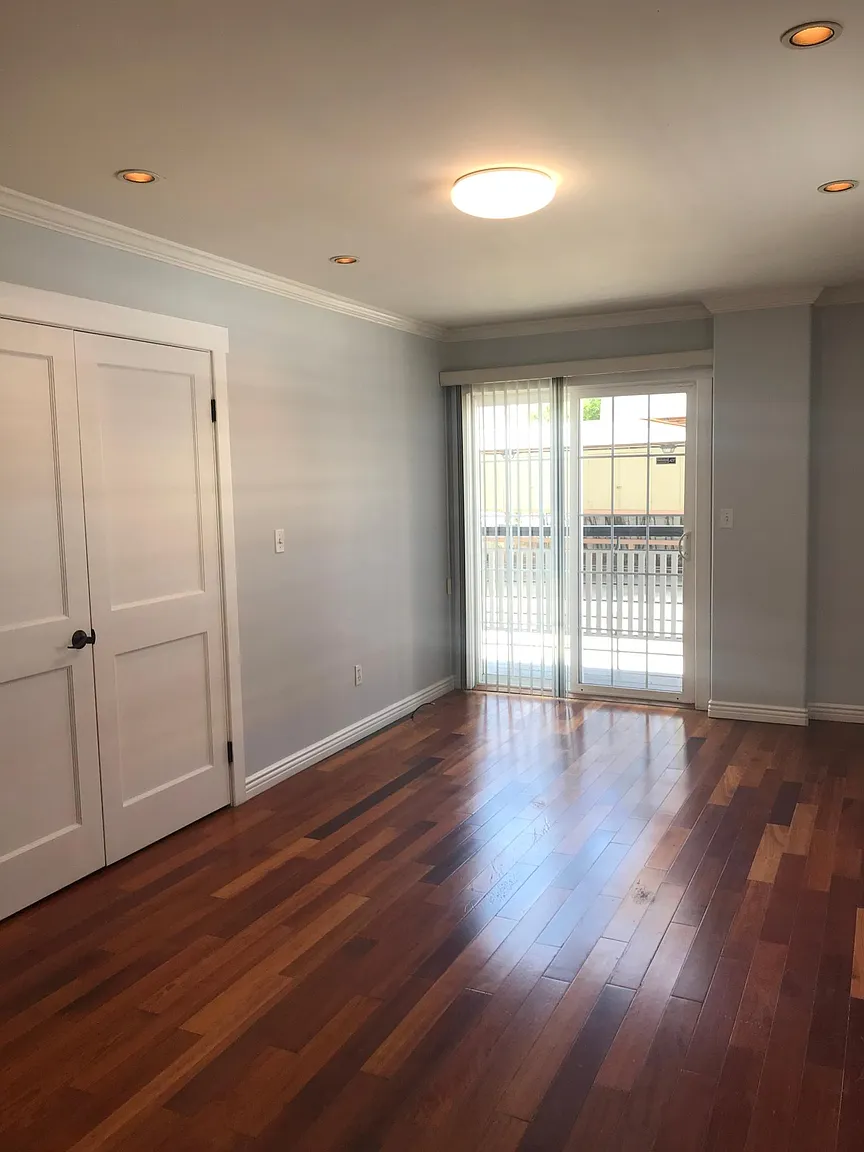 Empty room with hardwood floors, light gray walls, white baseboards, a closed double door, sliding glass door with vertical blinds, and ceiling lights.