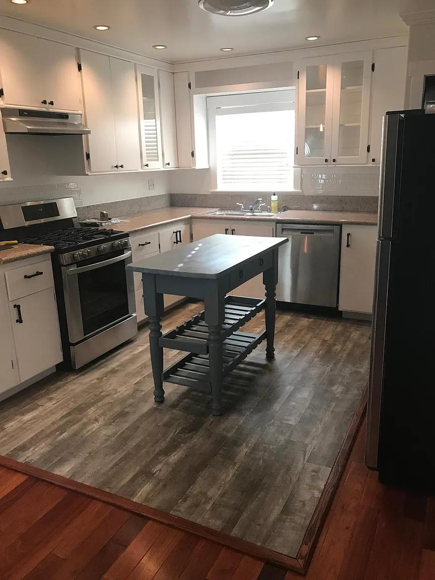 A kitchen with white cabinets, a gray countertop, a stainless steel stove, dishwasher, and refrigerator, a small gray kitchen island in the center, wood flooring, and a window above the sink with blinds.