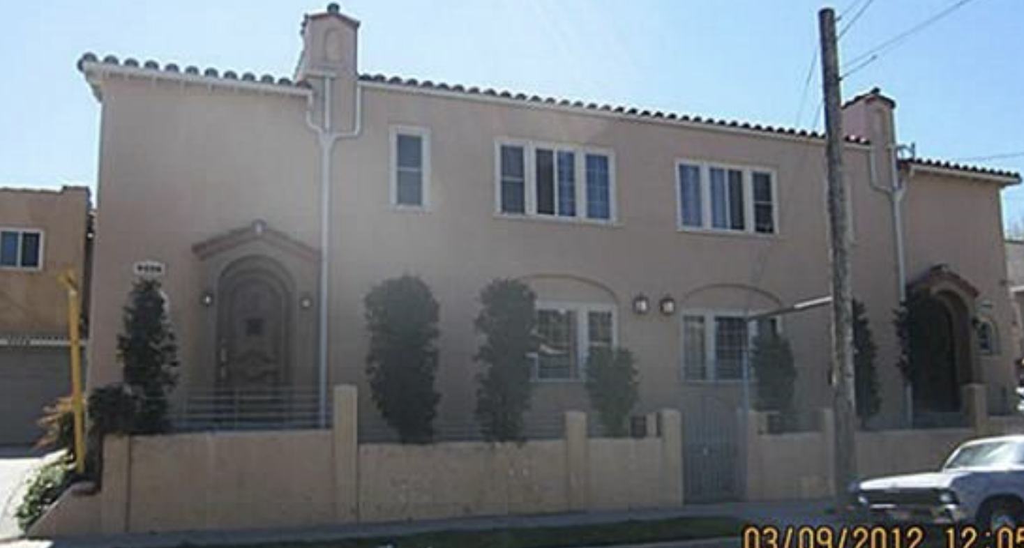 A two-story beige house with a tiled roof, arched doors, and several windows. There are three tall bushes in front of the house, a stone fence, and a utility pole nearby. A silver car is parked on the street.