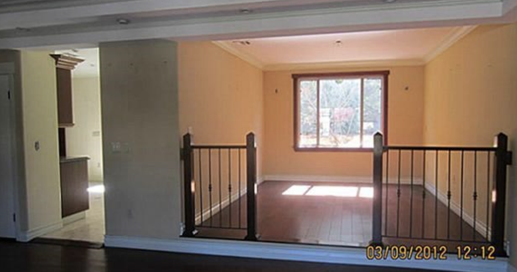 Interior view of a room with a wooden floor, large window, yellow walls, and a dark metal railing around a small sunlit area.