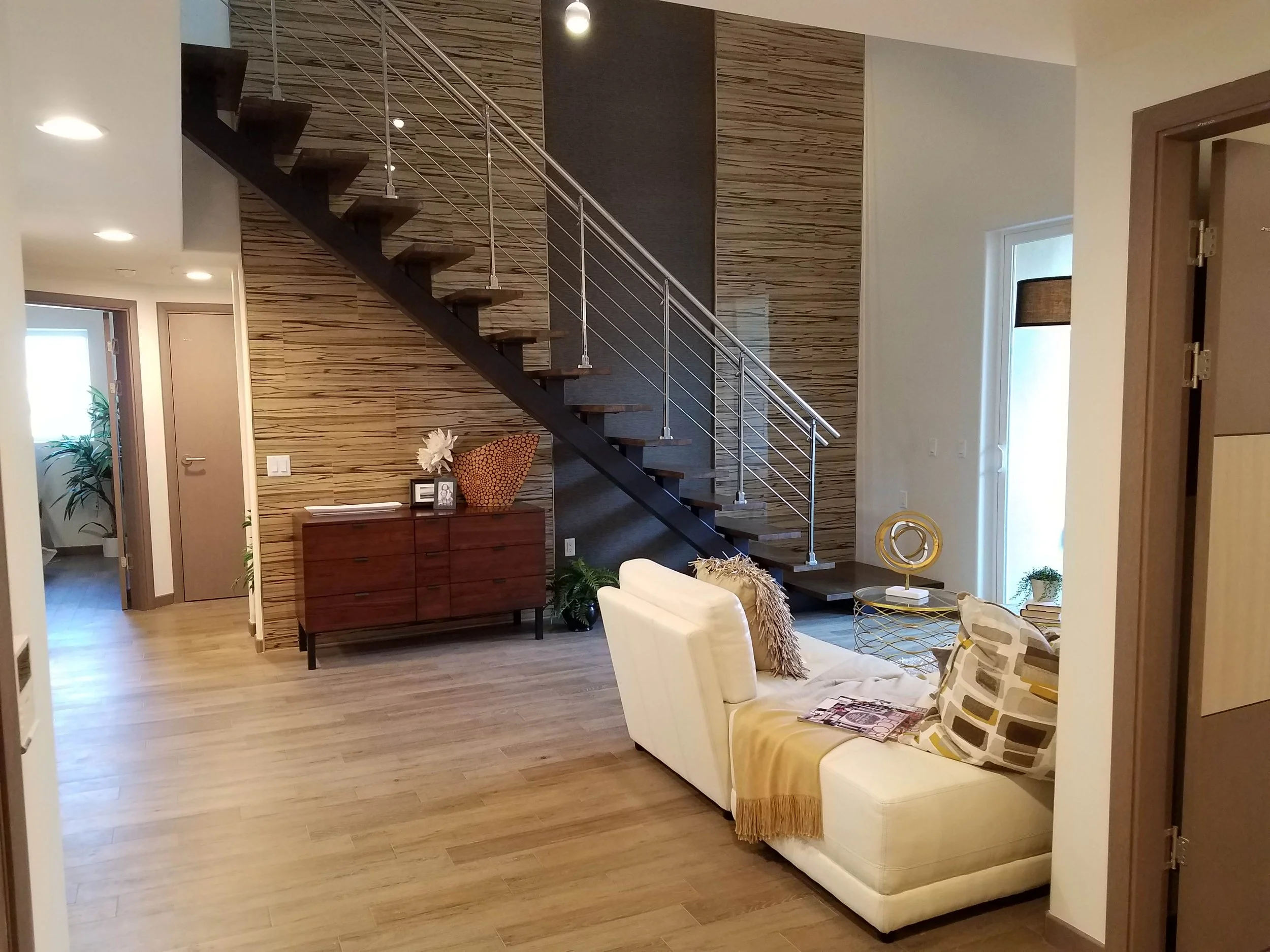 Living room with white sofa, decorative pillows, a yellow throw blanket, a gold coffee table, and modern decor, with a staircase featuring wooden steps and metal railing against a wood-textured accent wall.