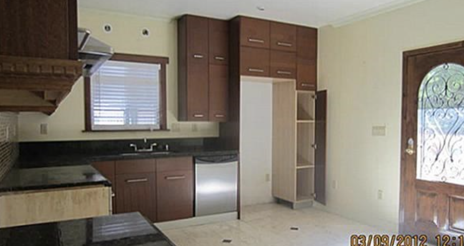 Empty kitchen with brown cabinets, black countertops, a small window with blinds, a door with intricate metalwork, and some missing or new appliances.