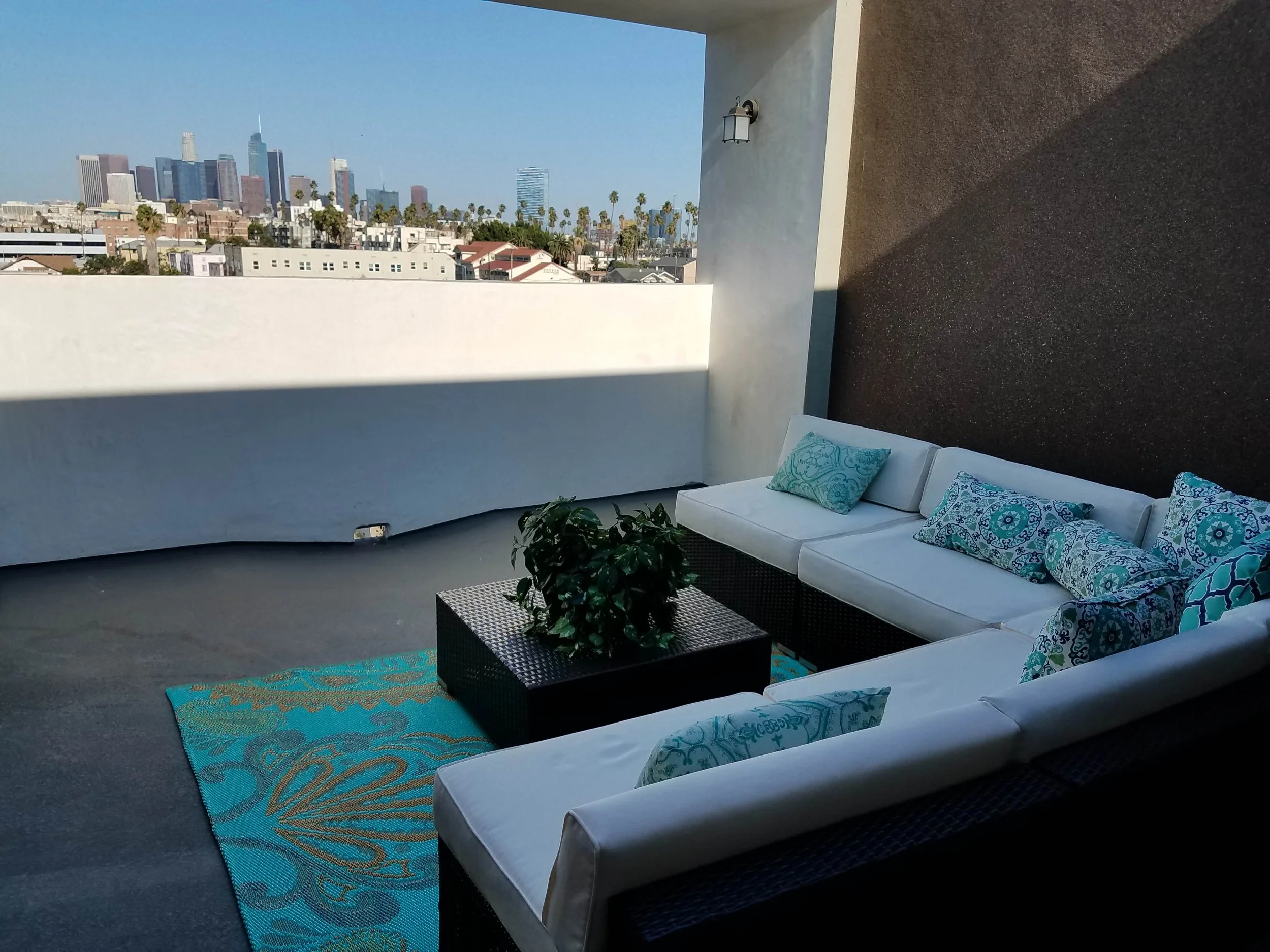 Outdoor rooftop patio with white sectional sofa and decorative pillows, potted plant on coffee table, city skyline in the background under a clear blue sky.