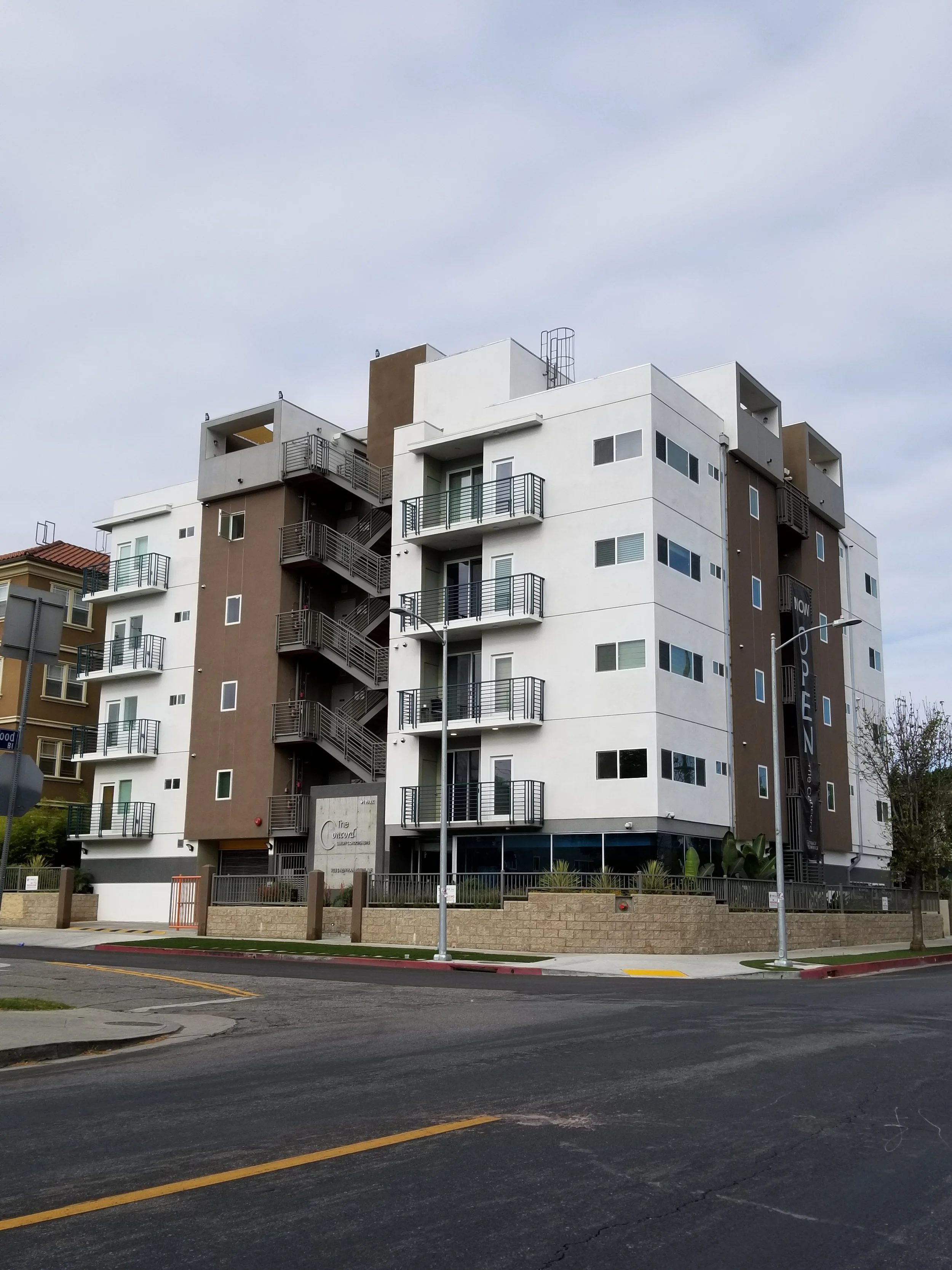 Multi-story modern apartment building with balconies and exterior stairs, located on a street corner under a cloudy sky.