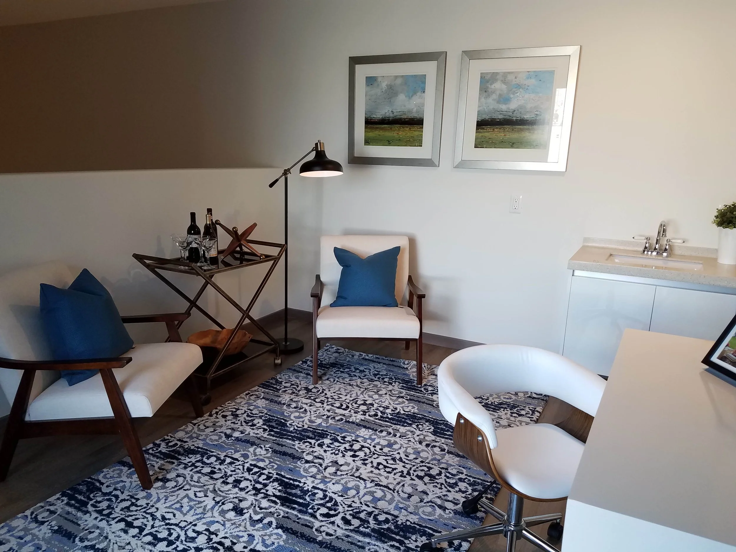 Living room with white and beige furniture, blue cushions, a patterned rug, a wall-mounted painting, a standing lamp, a small bar cart with drinks, a sink area, and a potted plant.