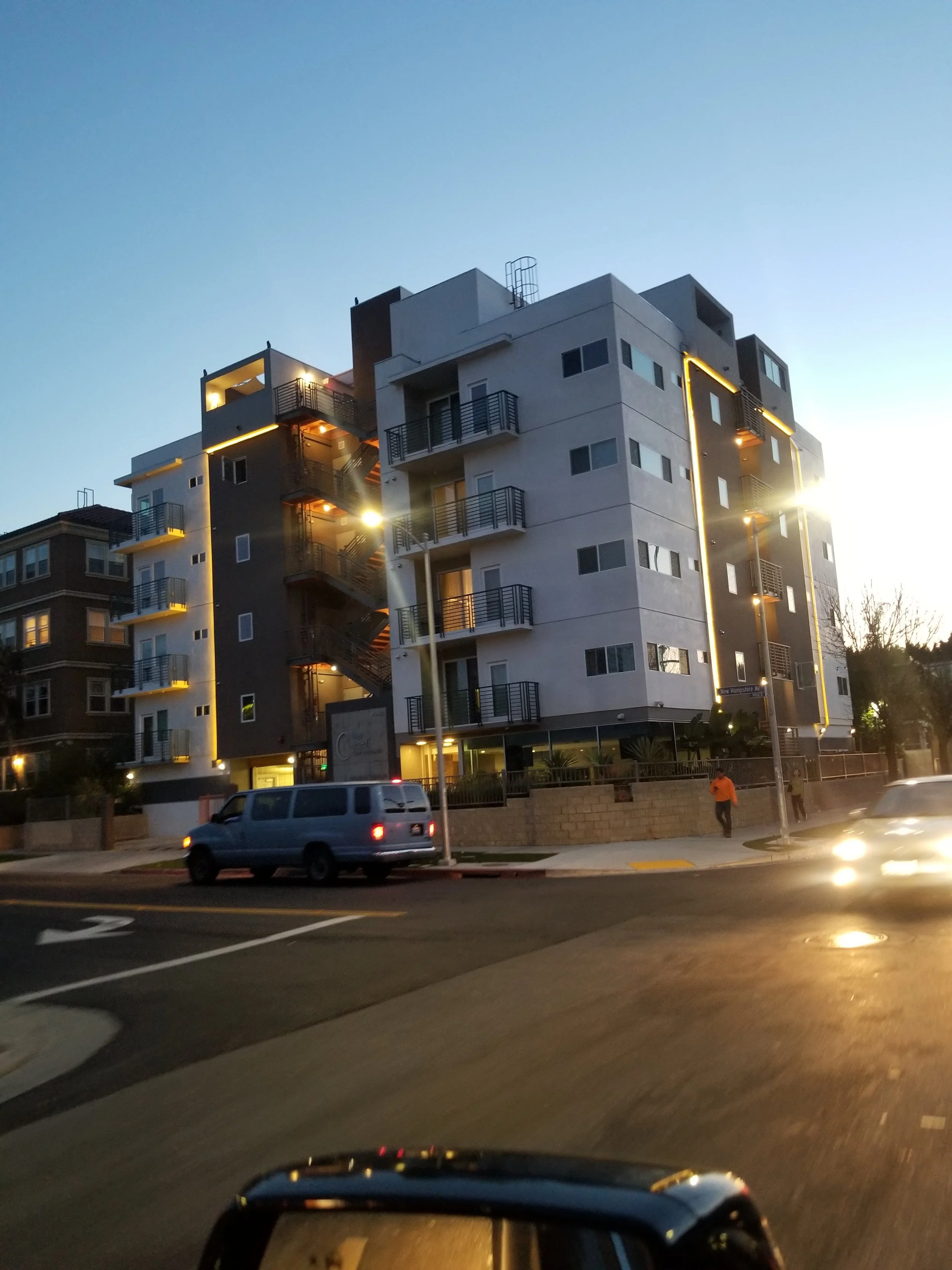 A modern apartment building with balconies and exterior lighting during dusk, with cars on the street and pedestrians nearby.