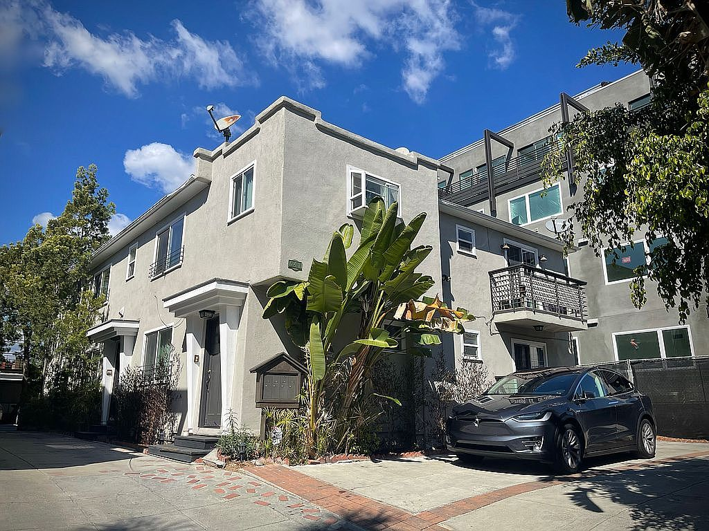 A modern multi-story apartment building with balconies, a black car parked in front, and lush greenery under a blue sky with clouds.