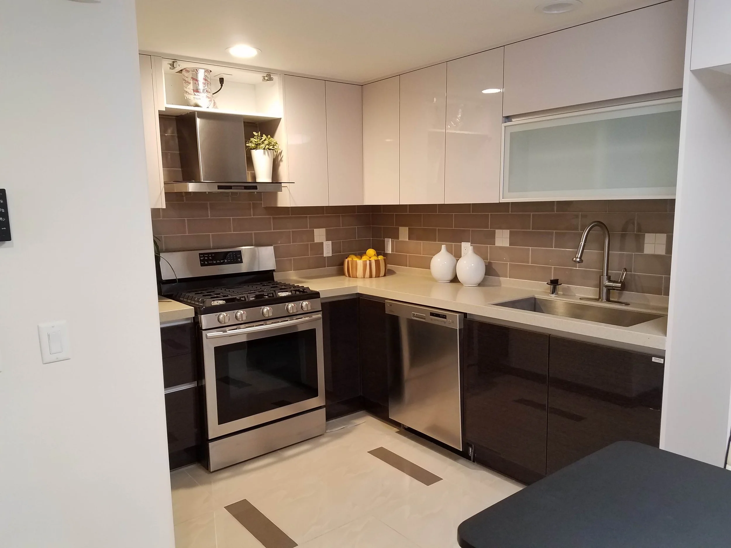 Modern kitchen with white upper cabinets, dark lower cabinets, beige countertops, stainless steel appliances, a brown tile backsplash, a stainless steel range hood, a stove, and a sink with a chrome faucet. Decorative vases and a bowl of lemons on the counter.
