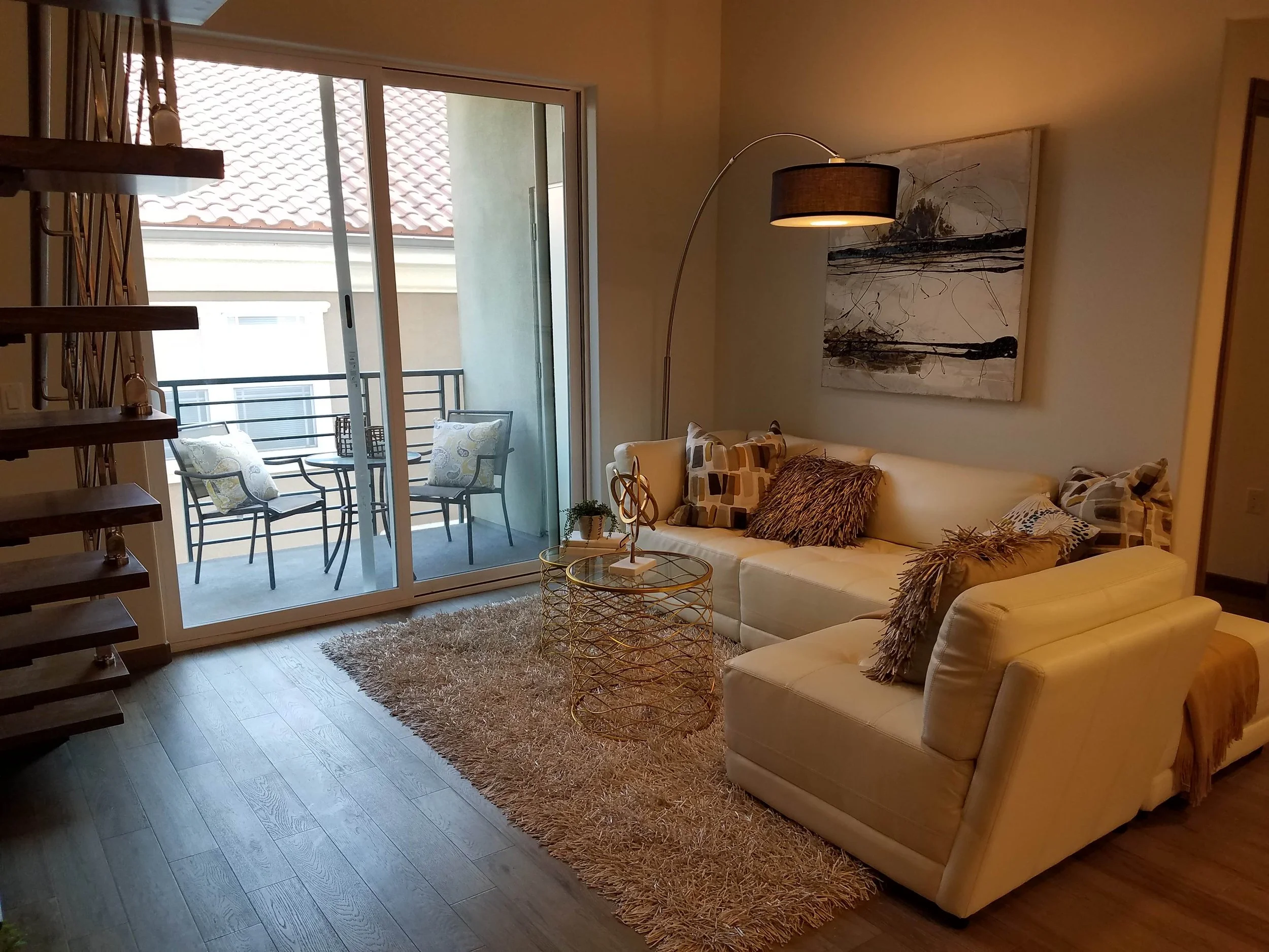 Living room with beige sofa, gold coffee table, and balcony with two chairs, all decorated in neutral tones.