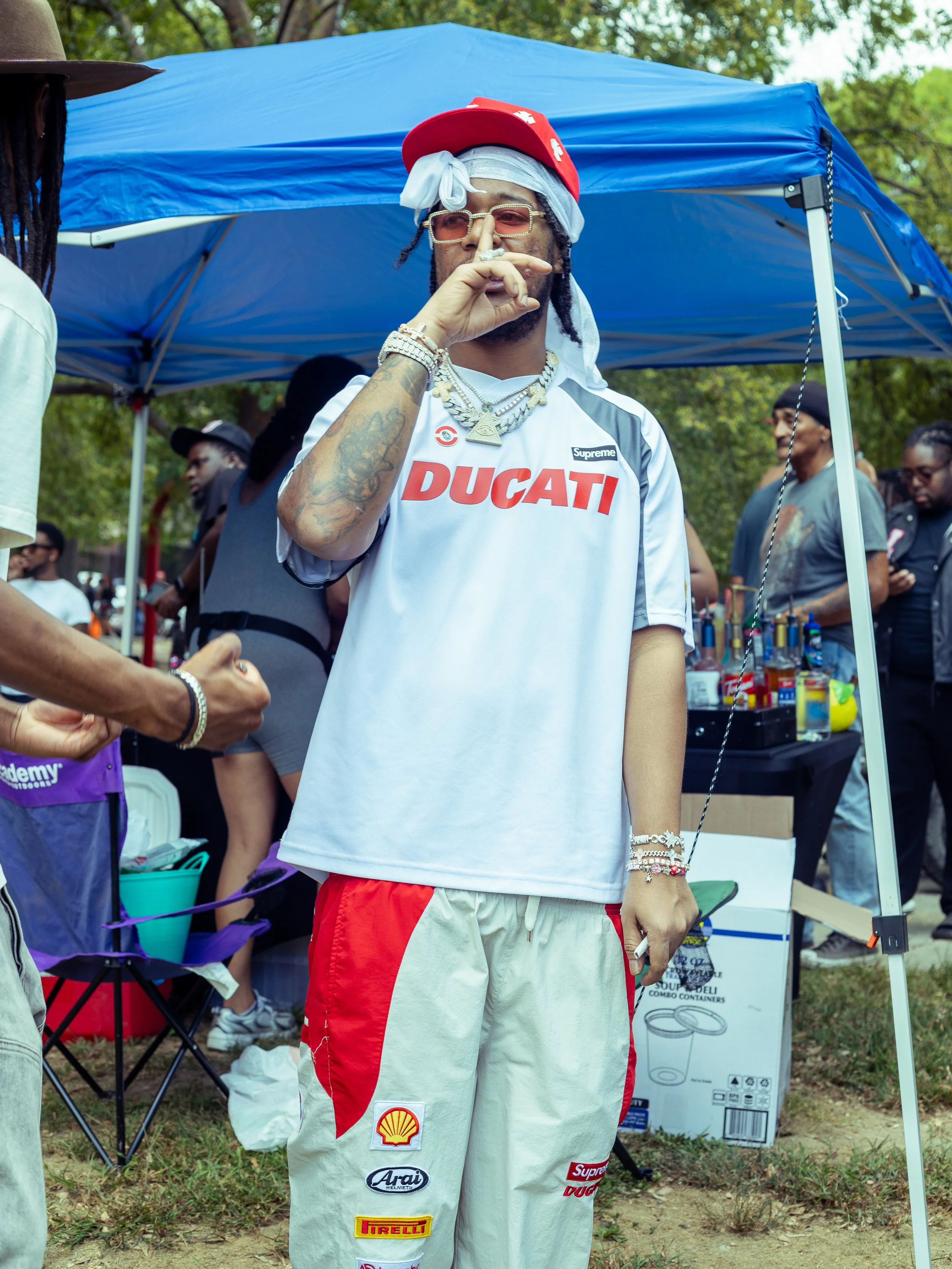 A man standing under a blue canopy at an outdoor event, wearing beige pants with various sponsor patches, a white racing jersey with Ducati and other logos, a red baseball cap, sunglasses, multiple necklaces, bracelets, and a bandana, holding a cigar